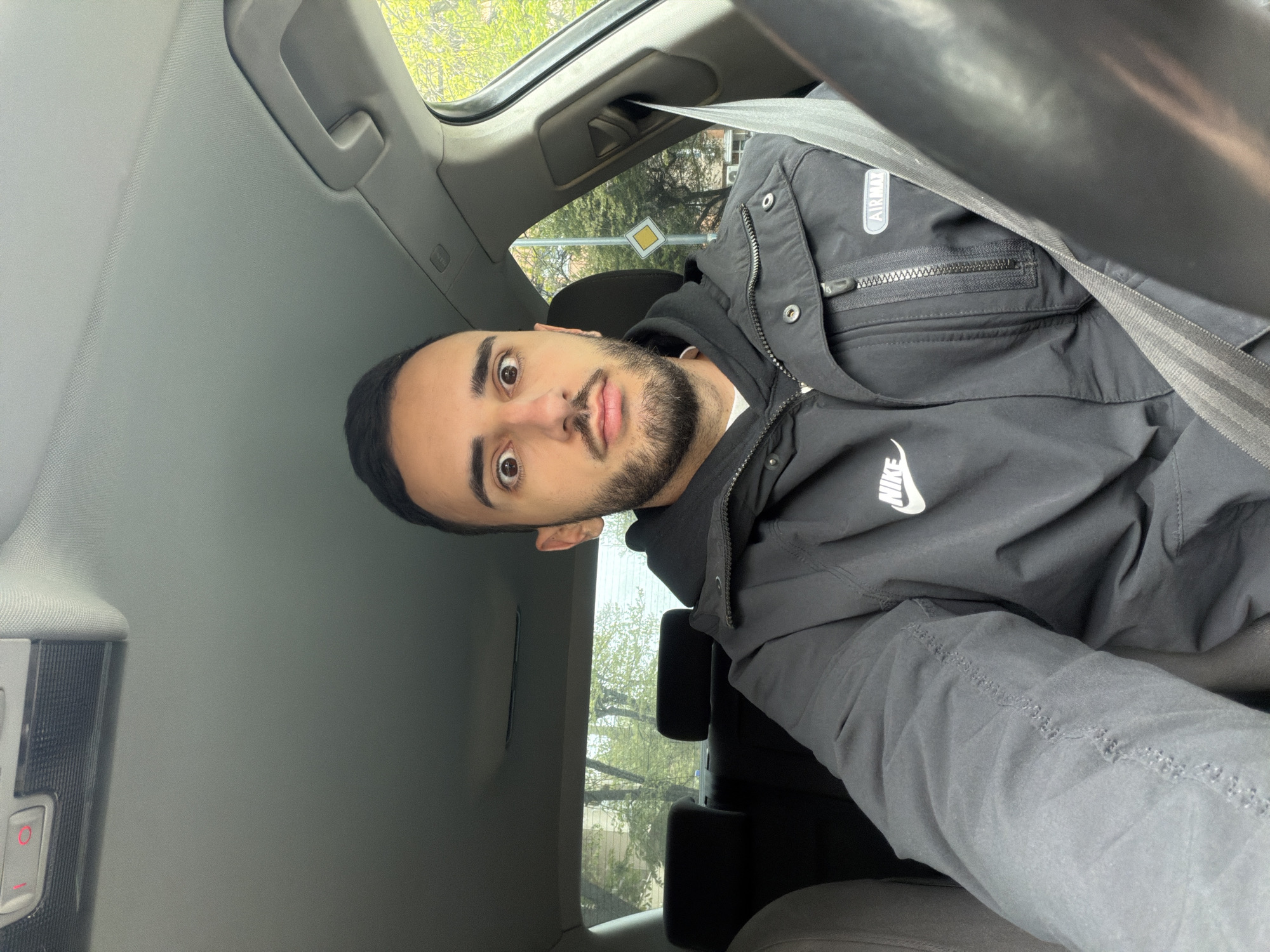 A man is seated in a car. He has dark hair, a beard, and brown eyes. He is wearing a black hooded sweatshirt under a black jacket with "Nike" and "Air Max" branding. A seatbelt is visible across his chest. Outside the car window, a street sign is visible, suggesting a location in an urban area, possibly Plovdiv, Bulgaria.