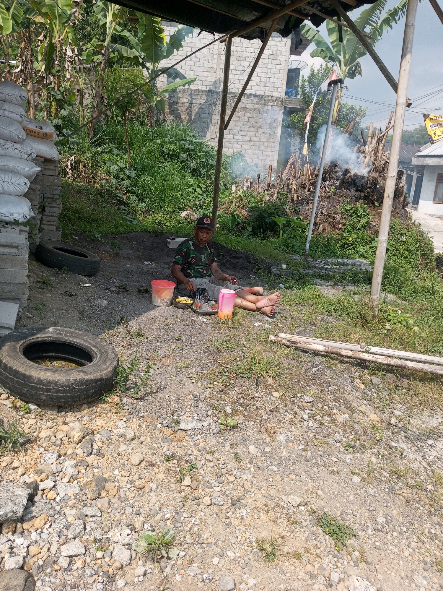 In Krasak, Indonesia, an elderly man in military-style camouflage clothing is seated on the ground under a makeshift shelter. He is accompanied by a few items, including a red plastic container, a metal bowl, and a pink plastic bottle. The ground is covered with dirt and gravel, with a discarded tire nearby. Behind the man, a pile of dried vegetation is smoldering, producing a thin plume of smoke. In the background, a brick building and lush vegetation can be seen.
