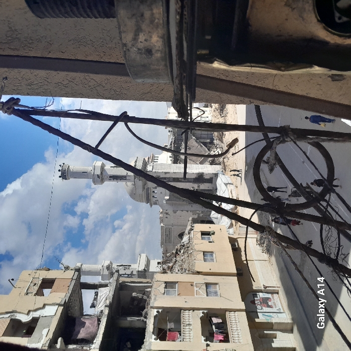 The image depicts a scene of extensive urban destruction in Khan Yunis, Palestinian Territory. In the immediate foreground on the right, a tangle of dark, corroded metal wires and rods, likely part of a damaged utility structure, partially frames the view. A portion of a textured, light-brown building facade is visible on the far right, along with a metallic cylindrical object mounted on a horizontal fixture.
A paved street or cleared pathway occupies the midground, where approximately eight to ten individuals are seen walking. One person in a blue jumpsuit is visible on the far right of the street, carrying a yellow bag. Other pedestrians, alone or in small groups, move further along the path towards the center of the frame.
The background is dominated by the Al-Fara Mosque, identifiable by its white dome and two prominent minarets with crowned tops, which appears structurally compromised with visible damage around its base. Surrounding the mosque are widespread areas of completely collapsed buildings, reduced to rubble and debris. White tarpaulins or tents are visible among these ruins. To the left of the mosque, a multi-story building exhibits severe structural damage, with entire sections of walls missing, exposed internal elements, and debris hanging. Laundry is visible drying on balconies of the partially intact sections of this building. The sky above is partly cloudy. The overall scene conveys a post-conflict urban landscape with residents navigating damaged infrastructure.