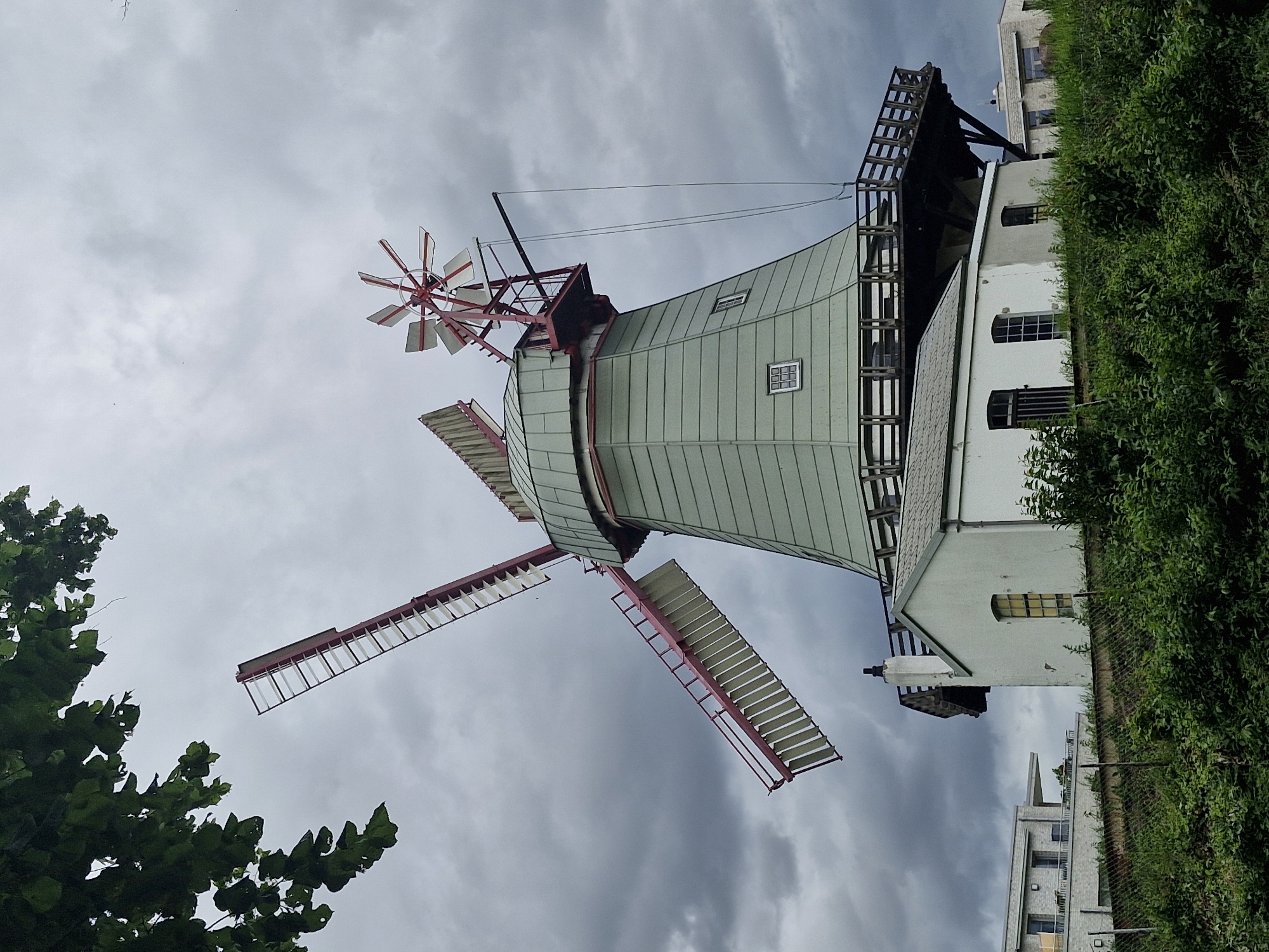An exterior, low-angle, medium shot captures a Dutch windmill situated in Bremen, Germany. The windmill has a conical, green-painted body with a red-and-white striped cap. One of its two large, red-and-white striped sails is extended. A small, white auxiliary building is located at the base of the windmill, with several windows. A wooden walkway with railings wraps around the windmill above the auxiliary building. Dense green foliage is visible in the foreground and upper left corner of the image. Modern buildings, one with a white brick facade and balconies, are visible in the background. The sky is overcast with dark clouds.