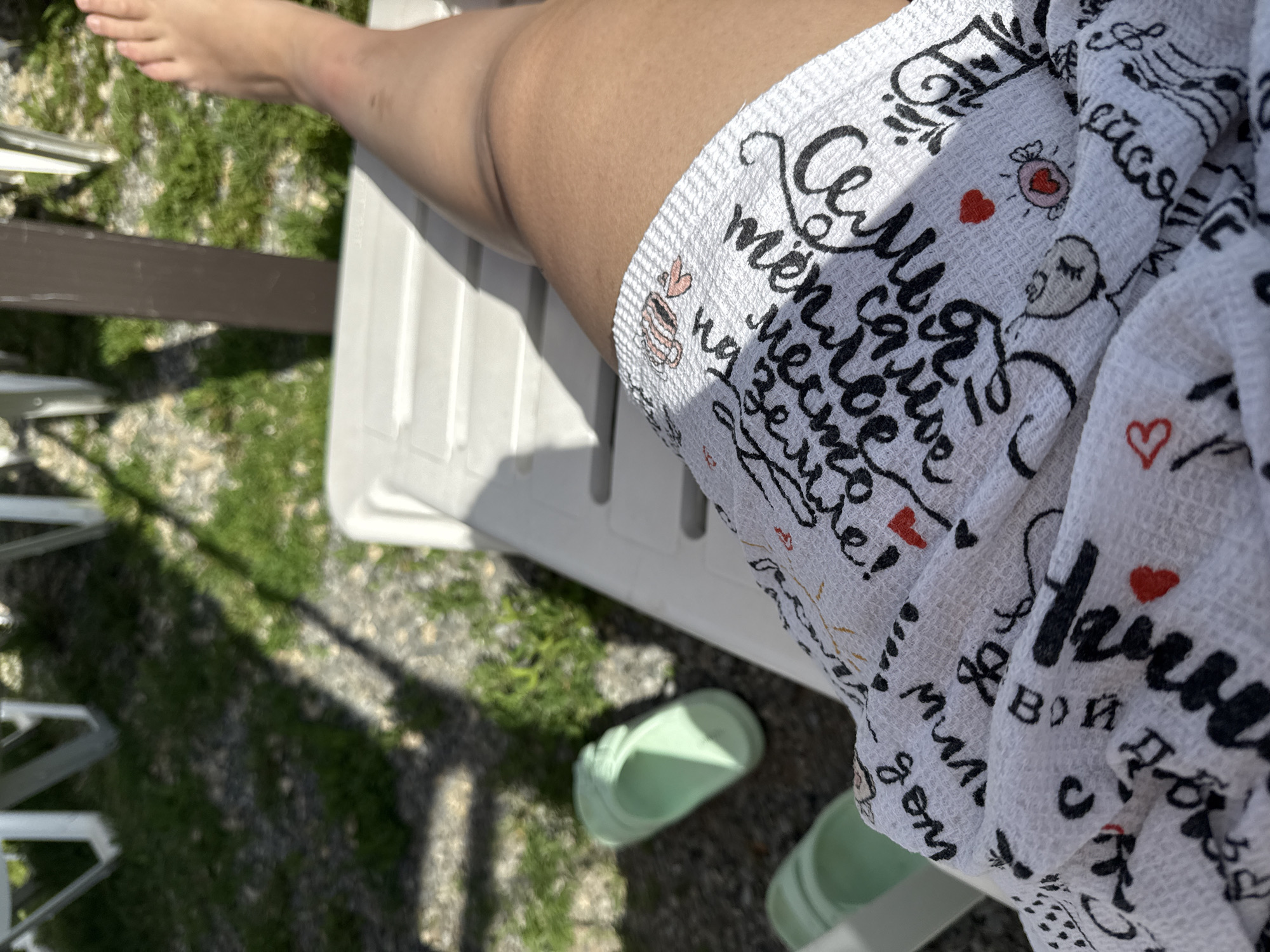 A person's leg and foot are visible, resting on a white lounge chair outdoors. The person is wearing shorts with a pattern of text and small heart and candy illustrations. Light green flip-flops are on the ground next to the chair. The background shows grass and the legs of other lounge chairs, suggesting a public outdoor seating area, possibly a park or resort. The lighting indicates it is daytime, with shadows cast by unseen objects.
