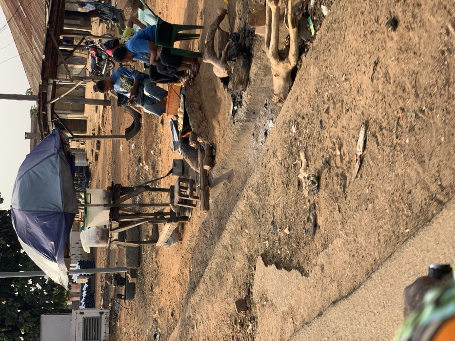 The image depicts an open-air, roadside scene in Kabba, Nigeria, characterized by an unpaved, reddish-brown dirt ground with a shallow drainage furrow running horizontally across the midground.
In the mid-ground, a makeshift food stall is present, covered by a blue and grey striped umbrella providing shade. The stall consists of a wooden frame holding two transparent plastic containers, one with a green lid, likely containing food items. A small wooden stool is positioned near the base of the stall.
Adjacent to the stall, to the right, three individuals are seated around an open-air cooking setup. An active fire, fueled by logs, burns beneath a metal pot or pan containing yellowish food items. A male, wearing a blue patterned t-shirt and jeans, sits on a light-colored plastic chair, looking down at and manipulating a mobile phone. To his right, a female, dressed in a blue t-shirt and dark trousers, is seated on a green plastic chair, observing the cooking area. A third person, partially obscured, is seated further to the right on another green plastic chair.
The ground around the cooking area and the drainage furrow contains scattered debris, including charred wood, ash, plastic litter, and a knife with a light-colored handle. In the background, multiple single-story buildings with corrugated metal roofs are visible, along with a parked motorcycle and a repurposed tire. To the far left, an external white unit and stacked pipes are visible. The scene is illuminated by natural daylight, casting subtle shadows.