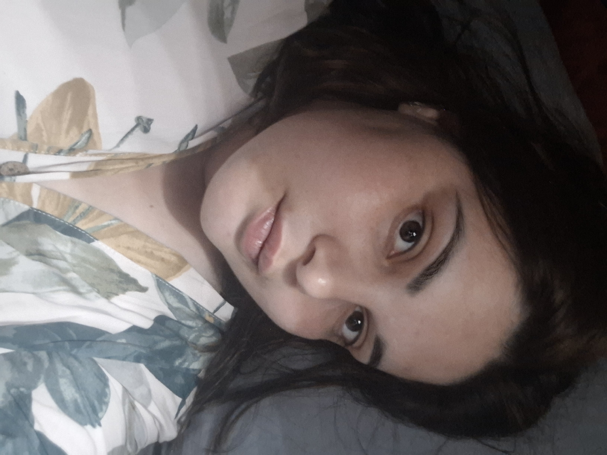 A woman with dark hair is lying down, her head tilted towards the left. She has fair skin and is wearing a patterned top with floral designs in shades of white, yellow, and blue. The background appears to be a pillow or bedding in a solid grey color. The photo seems to be taken indoors. There is no visible indication of the location being in the city of Амзя, Russia.
