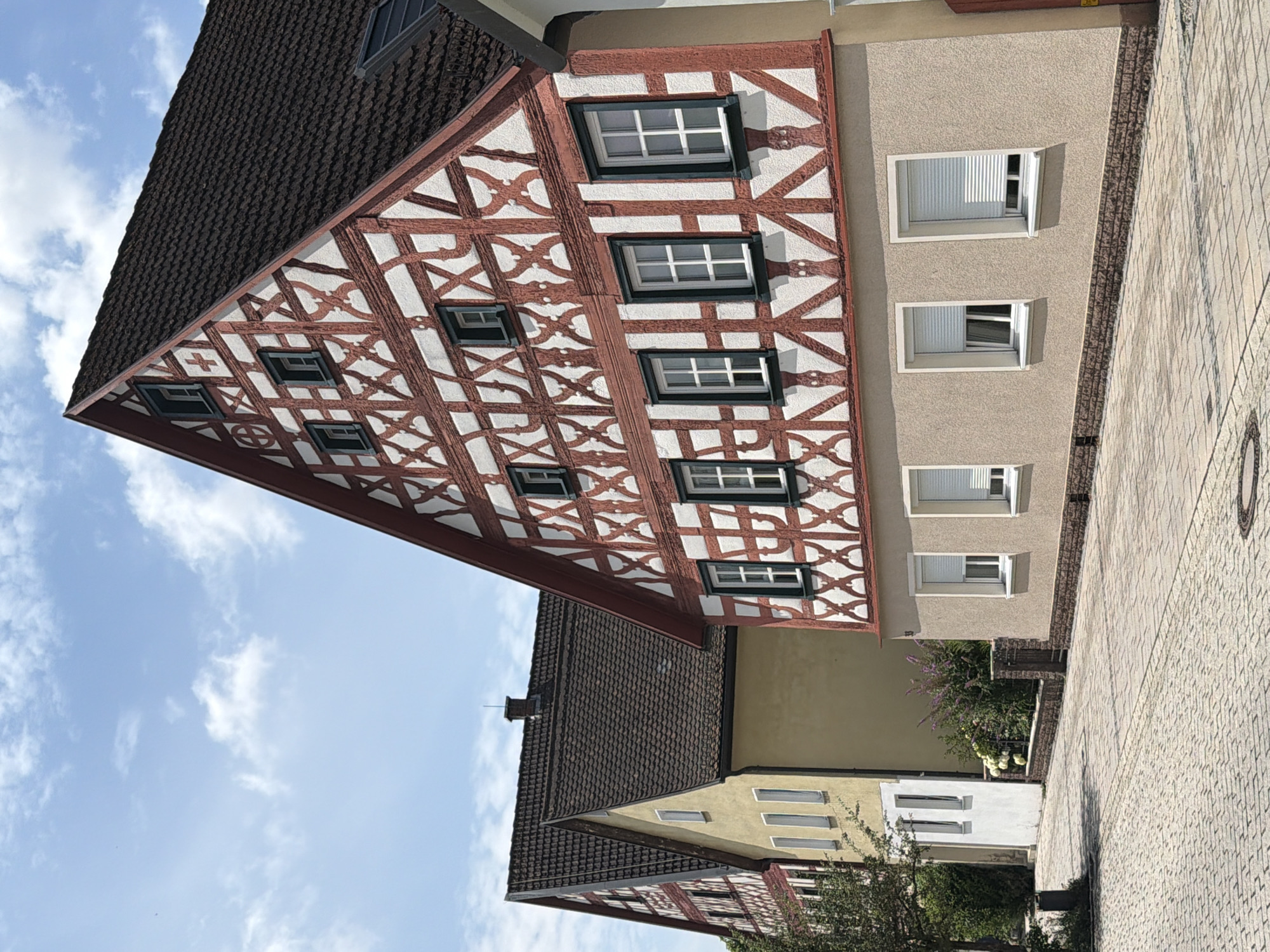 The image shows a view of traditional German timber-framed houses in Ebermannstadt, Germany. The house in the foreground features a prominent red and white timber-frame facade with multiple windows. To the left of this house, another timber-framed house is partially visible, followed by a yellow building with white trim. The street in front of the houses is paved with cobblestones, and there is a stone wall with some vegetation behind it. The sky is blue with scattered white clouds, indicating a daytime scene. There are no people visible in the image.