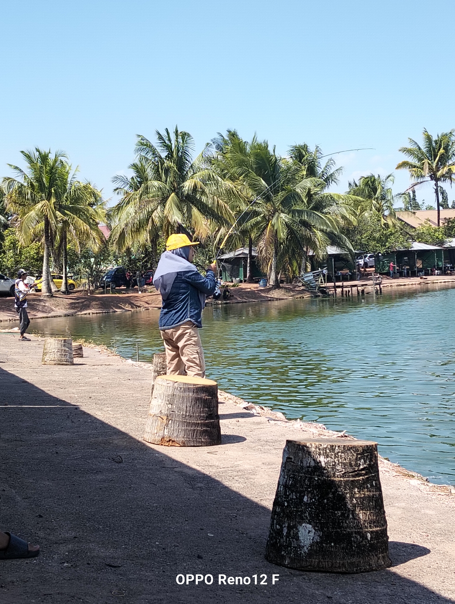The image depicts a fishing spot in Cindai Alus, Indonesia, with a person actively fishing. The foreground shows a concrete walkway with several tree stumps serving as natural seating. In the midground, a person wearing a yellow cap and a blue jacket is fishing with a rod and reel, facing away from the camera towards the water. Another individual, dressed in dark clothing with a white and red patterned shirt, is also fishing further down the walkway. The background reveals a serene lake surrounded by lush palm trees and several simple structures with green roofs, likely fishing stalls or small buildings. The sky is clear and blue, indicating a sunny day. The water appears calm and reflects the surrounding greenery. Several parked vehicles are visible near the structures, suggesting a recreational area. The overall scene conveys a relaxed atmosphere of leisure and outdoor activity.