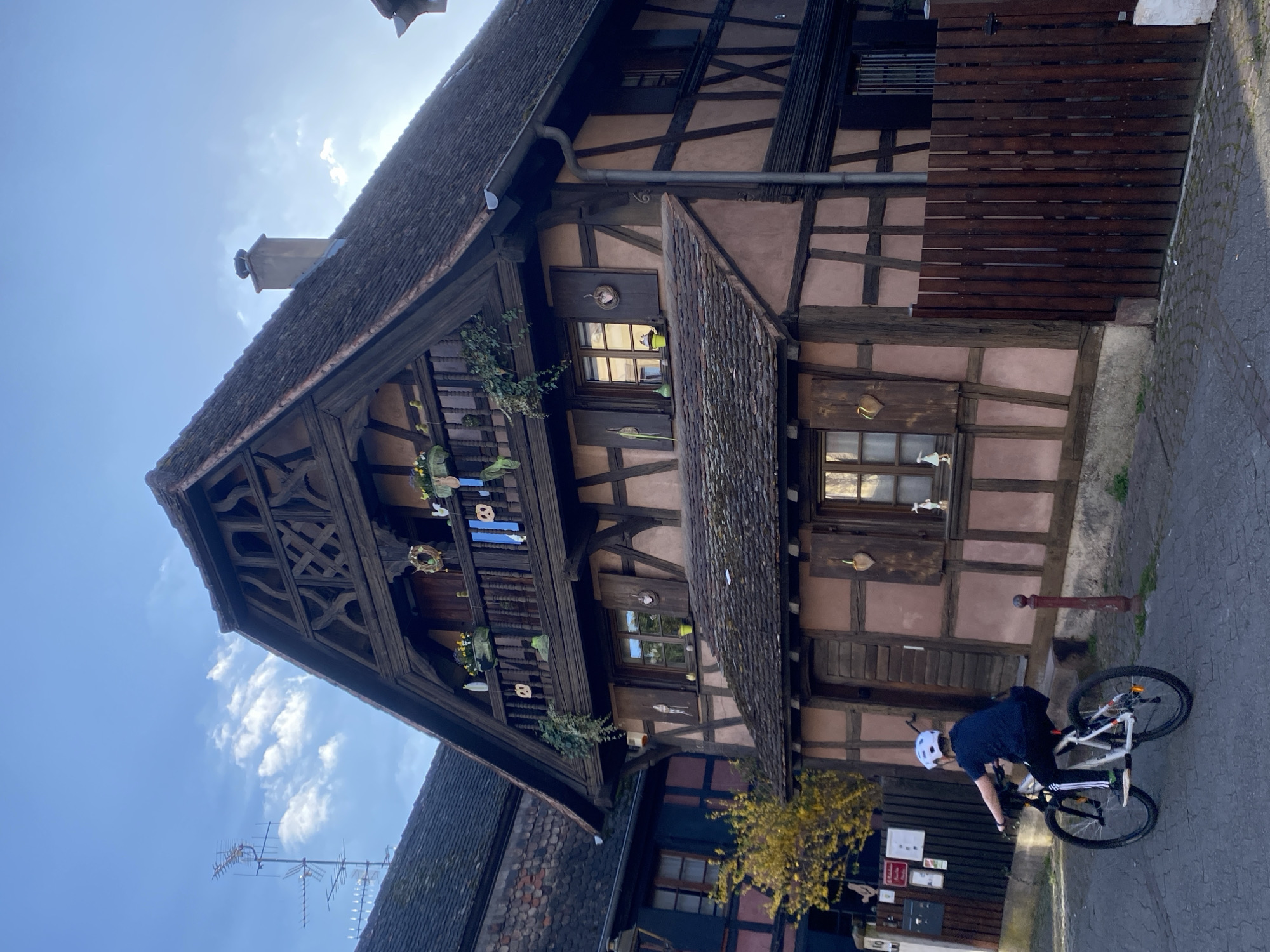The image depicts a person riding a bicycle on a cobblestone street in front of a half-timbered house. The house has a steeply pitched roof, multiple windows with shutters, and decorative elements on the balcony. The person is wearing a helmet and dark clothing. In the background, there is another building and a few plants. The sky is blue with some clouds.
Although not explicitly visible, given the architectural style and location, the building is likely located in Schiltigheim, France.