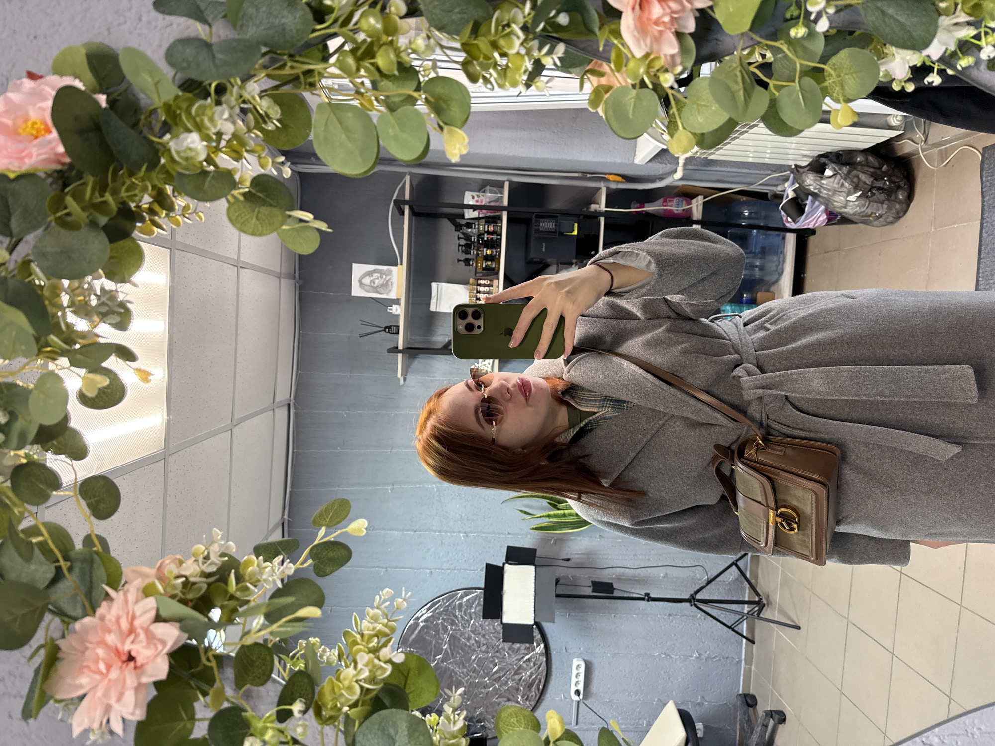 A woman with auburn hair stands in a room taking a selfie in a mirror. The mirror is surrounded by faux foliage, including eucalyptus leaves and pink flowers. The woman wears sunglasses and a gray coat with a belt. She has a small brown purse over her shoulder. The background shows a room with various equipment, including lighting stands, reflectors, and shelves with assorted items. The wall is painted a light blue-gray color. The tiled floor is a light color, possibly beige or white. Based on the room's appearance, it may be a beauty salon or photography studio in Chaikovsky, Russia.