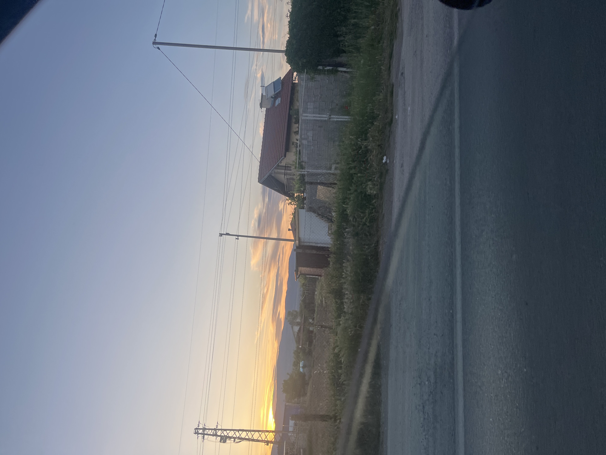 The image captures a scene in Blagoevgrad, Bulgaria during what appears to be sunset. The sky transitions from blue to orange near the horizon. Utility poles and power lines run parallel to the road. There is a building with a reddish roof and solar panels near the road, with some vegetation covering the area. The perspective suggests the image was taken from a moving vehicle, with a portion of the vehicle's interior visible on the right side.