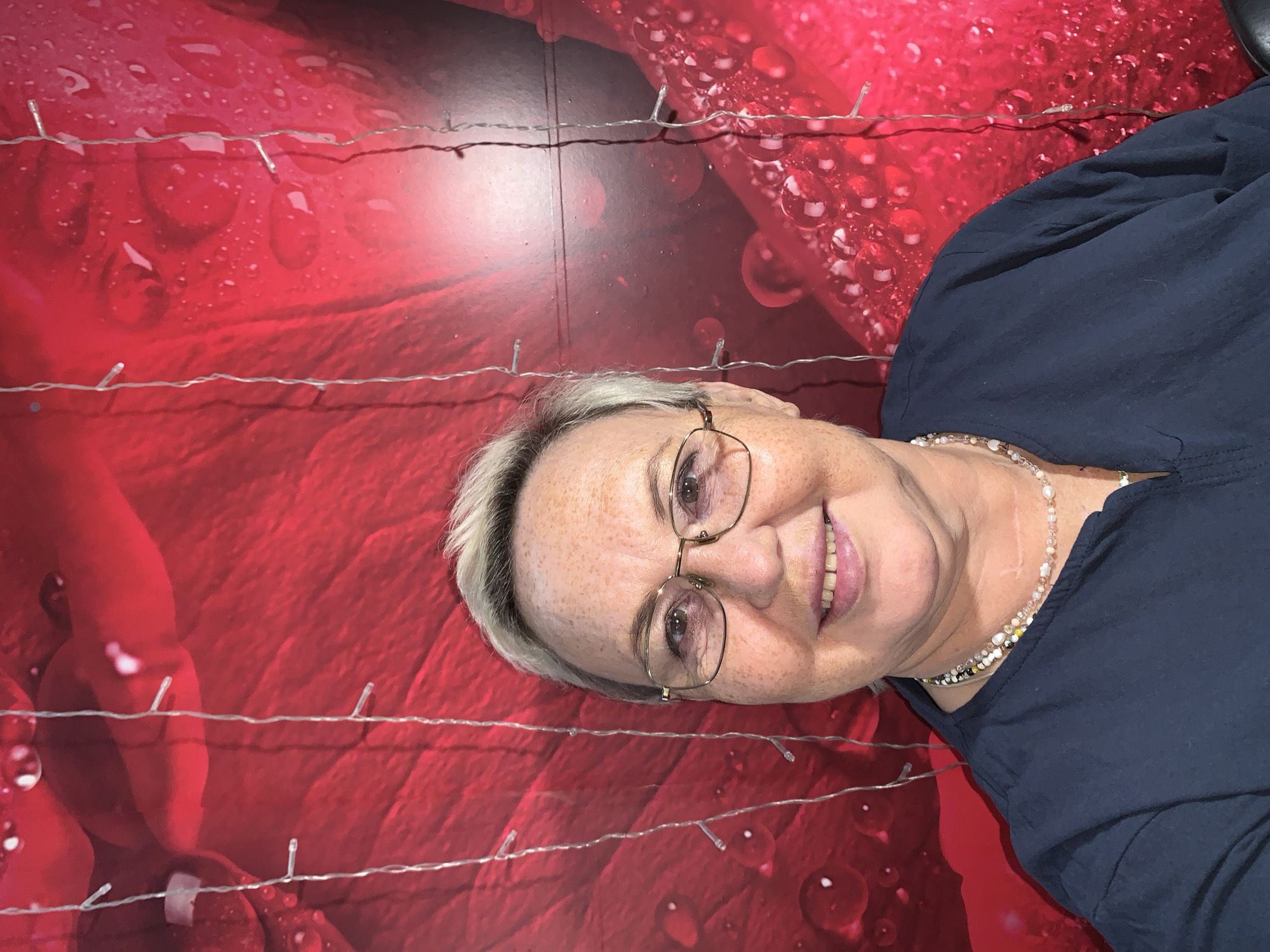 A middle-aged Caucasian woman with short, blonde hair and fair skin with freckles is looking directly at the camera with a slight smile. She is wearing glasses with gold frames, a dark blue shirt, and two layered necklaces. She is positioned in front of a bright red background that appears to be a fabric or wall covering with a pattern of water droplets. Several strands of small, white LED string lights are hanging vertically in front of the background, and some of them are in front of the woman. The woman is facing the camera in a straightforward manner, with her head and shoulders visible. The lighting is even, highlighting her features and the details of the background. There is no specific indication of the city of Chaikovsky, Russia, or any urban environment in the image. The scene appears to be a simple portrait setup.