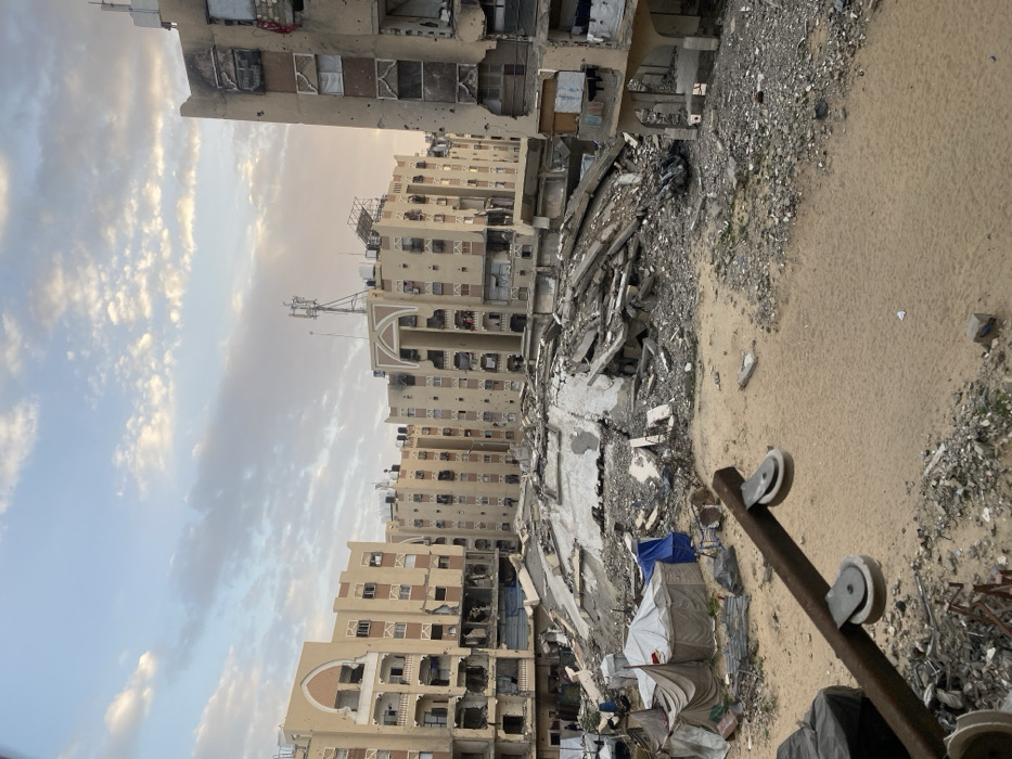 War-torn buildings dominate the skyline, pockmarked with damage and missing walls, revealing skeletal structures. Rubble and debris fill the space between the structures. Tents provide meager shelter amidst the devastation. A makeshift handcart sits foreground, hinting at resilience. The sky is soft with muted hues, a stark contrast to the grim reality below.