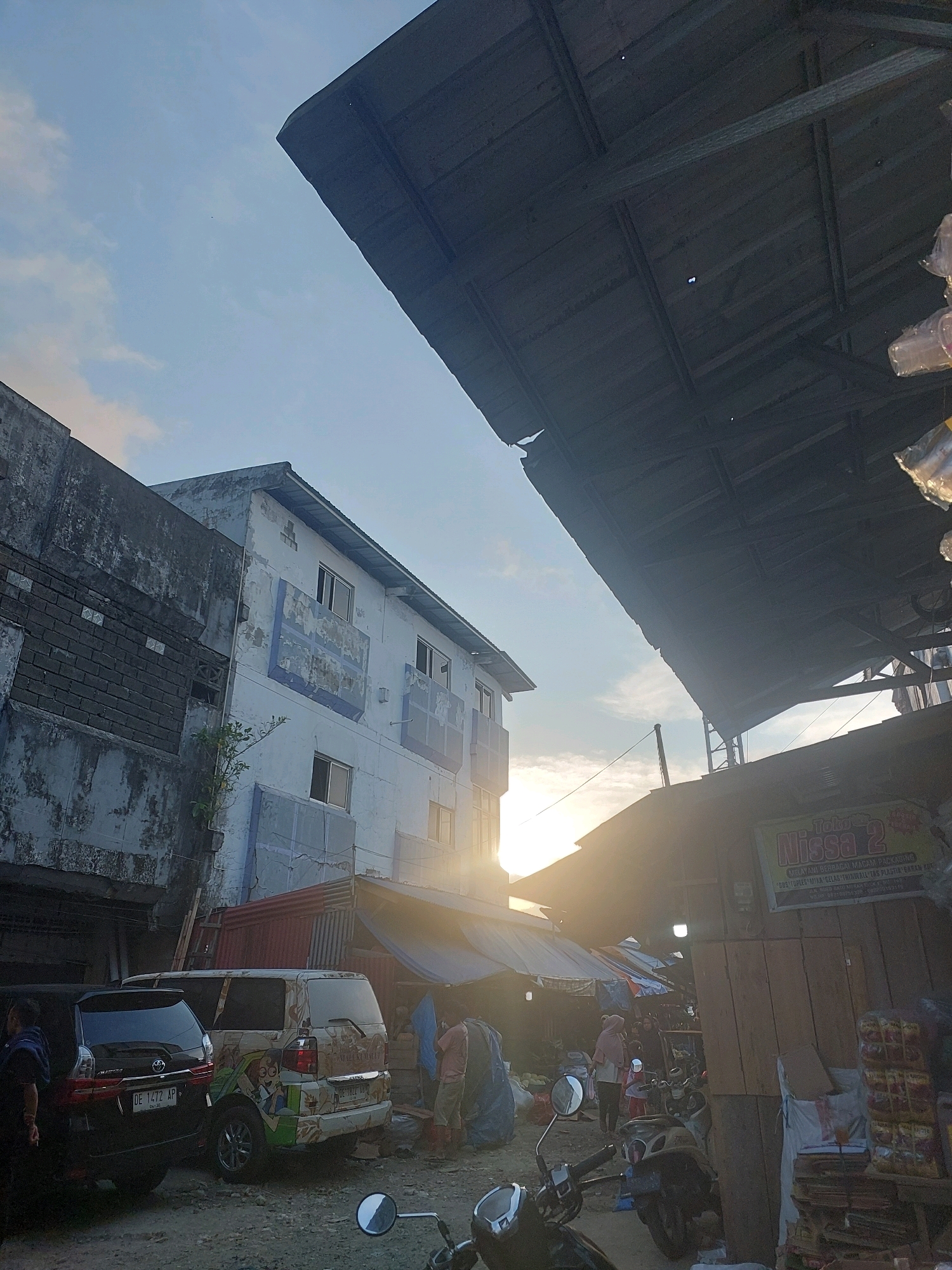 A low-angle, sun-drenched scene captures a street market in Ambon, Indonesia, with the setting sun casting a warm glow. In the foreground, a motorcycle's handlebars and a portion of its mirror are visible. To the left, two vans are parked. The first is a dark-colored van with license plate "DE 1472 AP". Next to it is a lighter-colored van adorned with cartoonish decals.
Behind the vans, a multi-story building stands, its white facade weathered and peeling, with blue decorative elements. Adjacent to it, a darker, rougher wall shows exposed brickwork. Further into the scene, a sprawling market unfolds under a rustic, dark-colored roof structure. Stalls are set up, with awnings and tarps providing cover. Several people are present in the market, some appearing to be vendors or shoppers. Goods are visible in the stalls and piled on wooden structures, including what appear to be stacks of packaged food items on the right. A sign for "Toko Nissa 2" is affixed to a wooden structure within the market. The overall impression is of a bustling, everyday scene at a local market in Ambon.