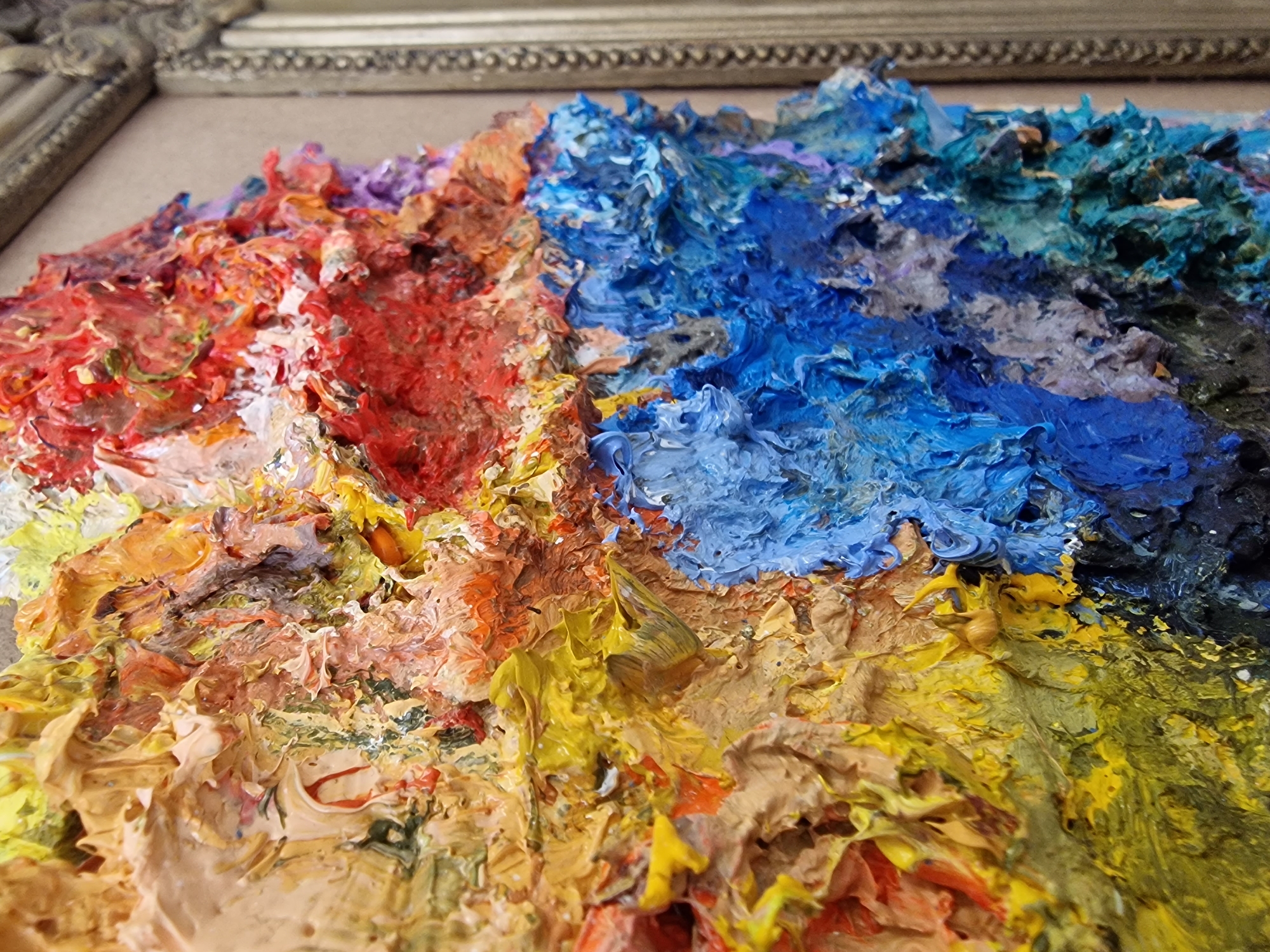 The image displays a close-up, oblique perspective of a heavily textured impasto oil painting. The artwork is characterized by an extremely thick application of paint, creating prominent three-dimensional peaks and valleys across its surface. Dominant colors include vibrant blues, rich reds, and various shades of yellow and orange, intermixed to form an abstract or impressionistic landscape. A segment of an ornate, possibly gilded, frame is visible along the right edge, framing the canvas. No individuals are present, and no specific actions or interactions are depicted. The scene is located in the city of Veliko Tarnovo, Bulgaria.