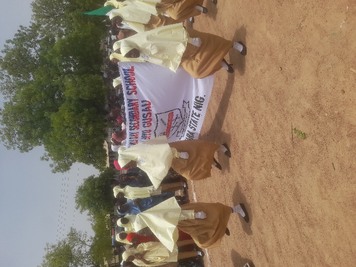 A group of people, possibly students, dressed in matching cream-colored head coverings and tops over brown skirts, are parading. They are carrying a white banner that reads "GOVT. DAY SECONDARY SCHOOL BIRNIN YERO GUSAU," with "ZAMFARA STATE NIG." below. The banner features a simple logo with a book. The background consists of a large green tree and a light brown dirt ground. The photo appears to be taken outdoors during the day.