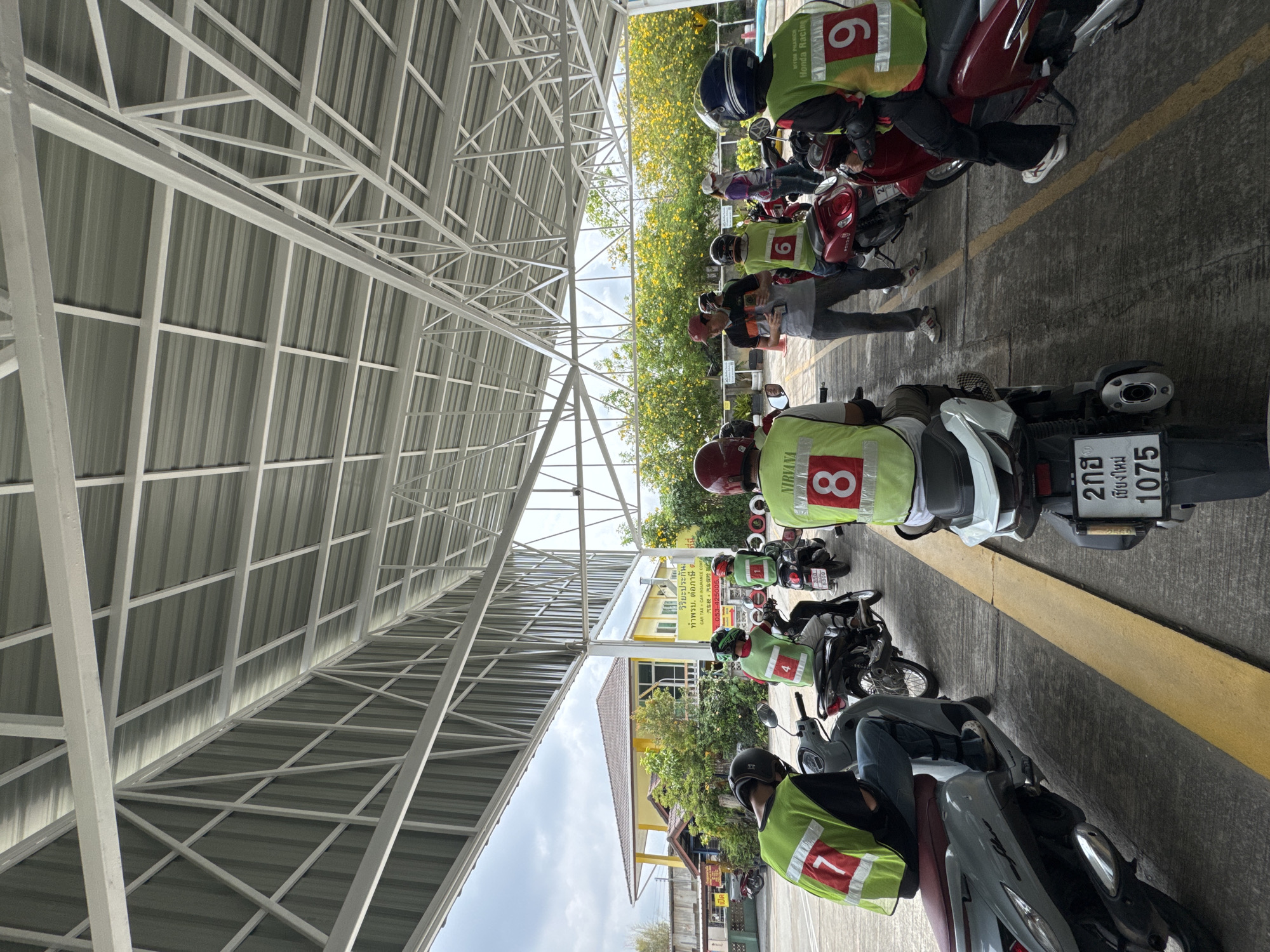 The image depicts a motorcycle parking area, likely in Nong Phueng, Thailand. Several motorcycles are parked in a row under a metal roof structure. Riders wear green vests with numbers on them, likely indicating a training course. One bike has the license plate "2กฮ เชียงใหม่ 1075". A building with a sign is partially visible in the background with text, "วิริยะประกัน ทำพรบ. ต่อภาษีด CAR TAX CAR INSURANCE CONTA". The area seems to be near a building providing car tax and insurance services. Some yellow flowering plants are also visible in the background.