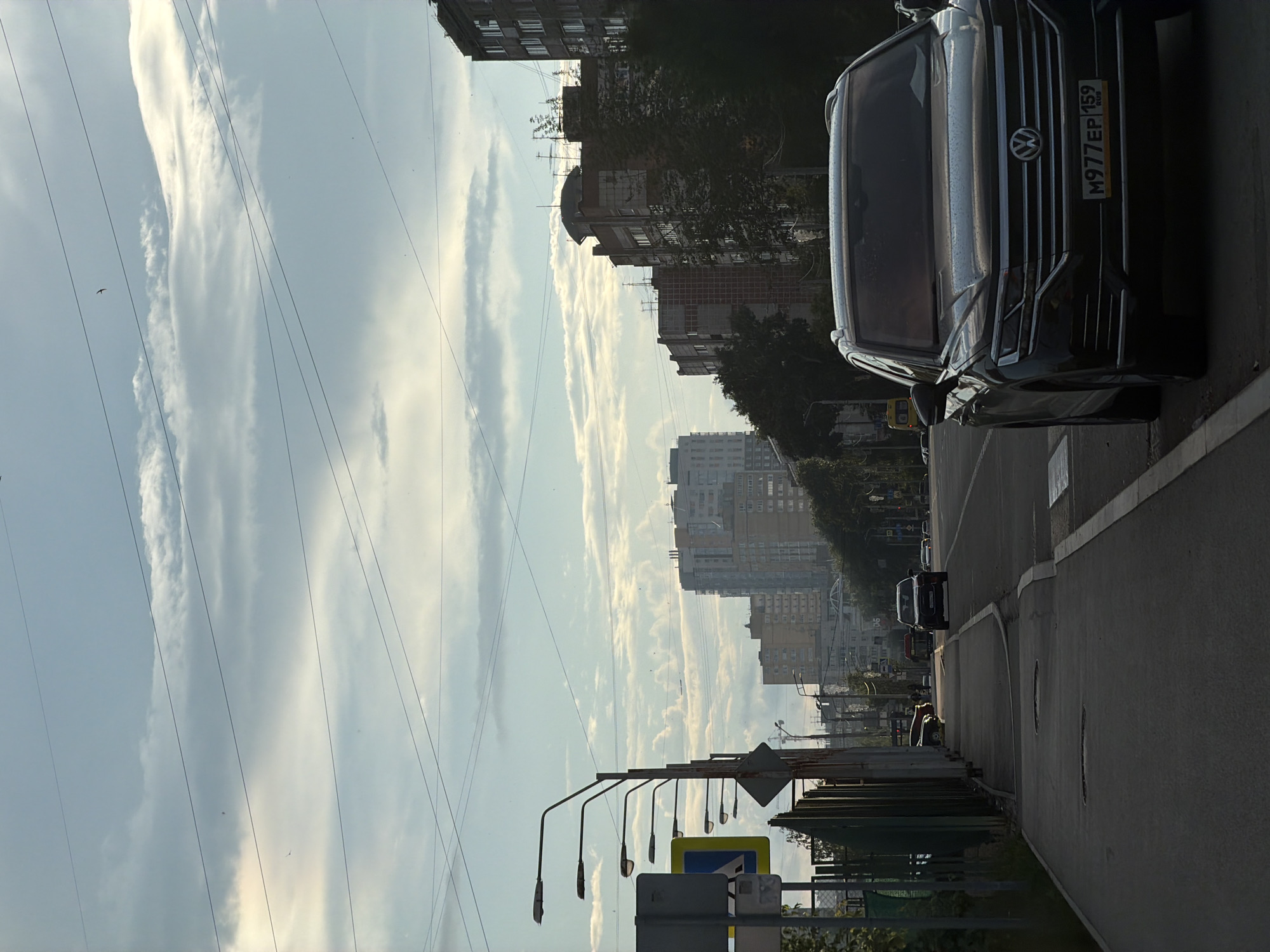 The image depicts a street scene in Perm, Russia, under a cloudy sky with some visible power lines. In the foreground, a dark-colored Volkswagen Touareg is parked on the right side of the road, facing forward, with its license plate reading "M977EP 159 RUS". To the left of the road, there is a sidewalk and a green fence. Further down the street, several cars and buses are visible, and tall apartment buildings line the horizon. The overall impression is that of an urban environment on a partly cloudy day.