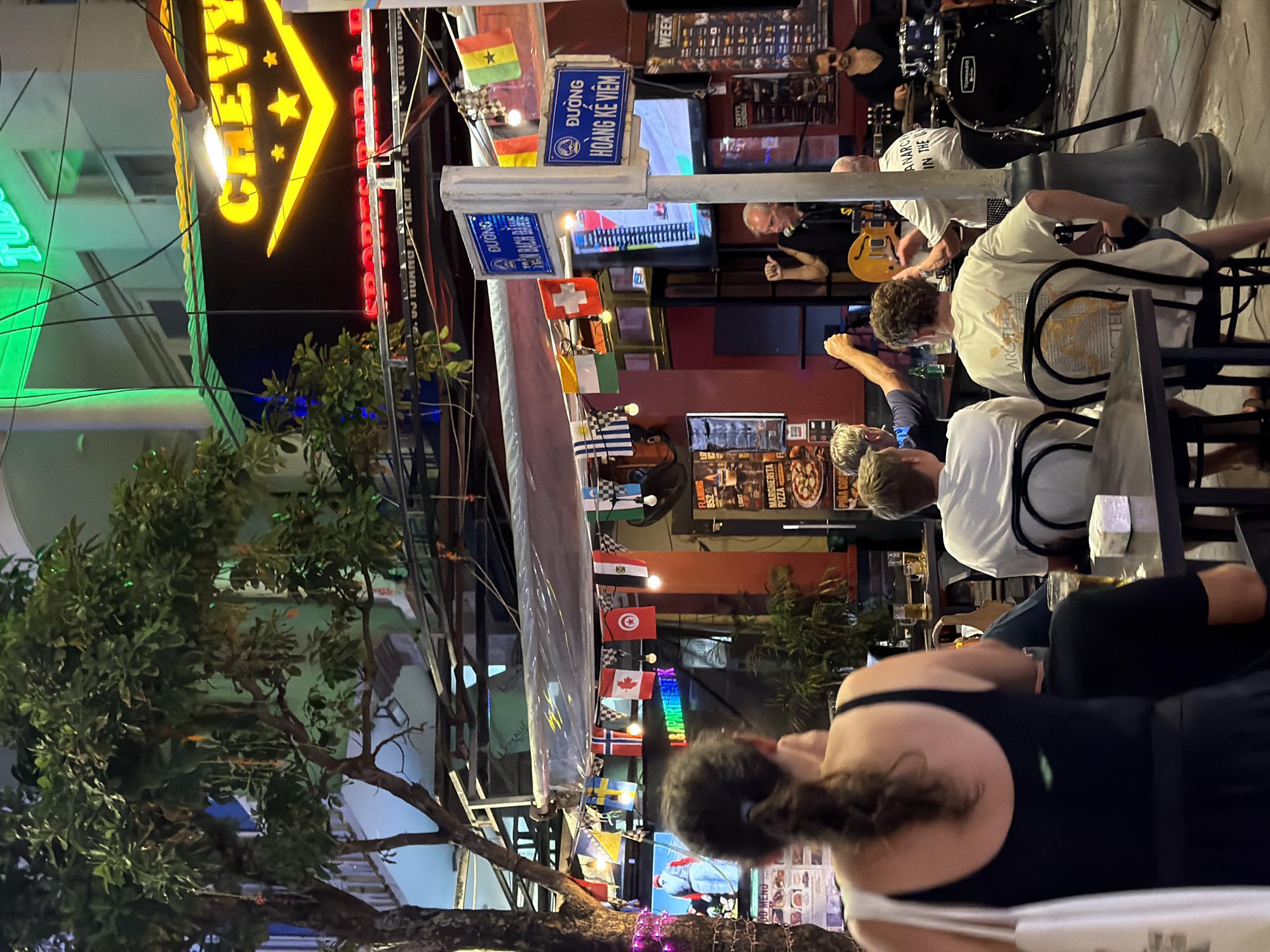 The image depicts an outdoor street scene at night in Da Nang, Vietnam, likely in an entertainment district. A neon-lit sign for "CHEVY" with a V-shaped emblem and stars is prominently displayed above a bar or restaurant. String lights with small flags of various countries, including Sweden, Norway, Canada, Tunisia, and Switzerland, are hung across the street. A live band is performing on a raised platform, with a guitarist and a drummer visible. Several patrons are seated at tables, some facing the band and others with their backs to the camera. A woman in the foreground is facing away, holding a white tote bag. Two street signs with Vietnamese text are mounted on a pole. The overall atmosphere suggests a lively, casual evening out.