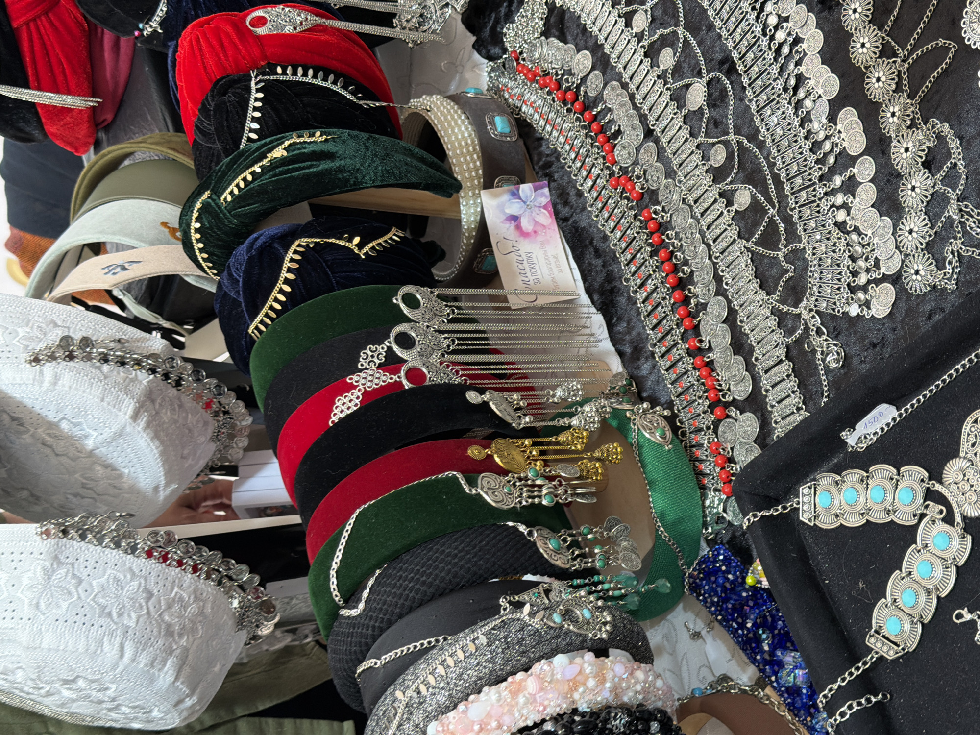 The image shows a close-up, high-angle view of a collection of handcrafted jewelry and accessories displayed on a black fabric surface and a wooden stand. The items appear to be of ethnic or bohemian style. In the foreground, several layered silver necklaces and waist chains are visible, featuring intricate metalwork, red beads, and numerous small coin-like charms. Some of these chains have a price tag indicating "1500". To the left, a black velvet tray holds more jewelry, including a necklace with multiple turquoise-colored stones. Behind these items, a series of velvet headbands are arranged, in colors such as black, green, red, and dark blue. These headbands are adorned with various embellishments including chains, gold-colored dangling elements, and what appear to be large, ornate silver earrings. At the top of the image, two white, lace-like hats or caps are prominently displayed, decorated with silver chains and small red and silver charms. A small card with floral imagery and Cyrillic text is placed among the headbands. The overall impression is of a market stall or display of artisanal goods. The lighting is even and there is no indication of the specific location or environment beyond the display itself.