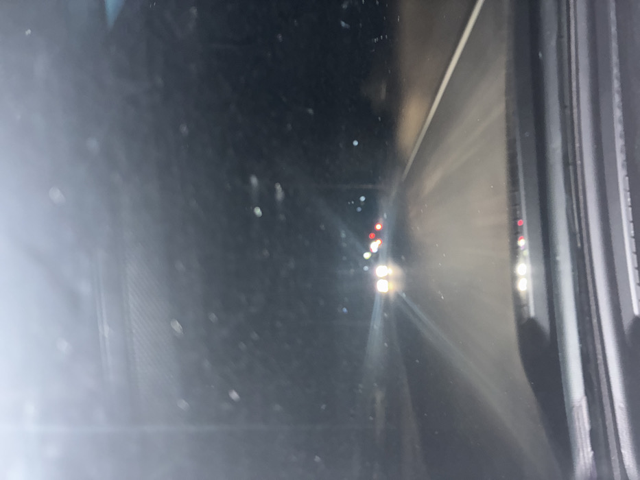 A nighttime view from a car's side mirror shows a highway receding into the distance. Bright headlights and taillights of vehicles punctuate the darkness. Streaks of light from oncoming cars create a hazy glow on the road surface. The side mirror frame is visible on the right side, with a blurred reflection on the left. The scene conveys a sense of speed and the isolation of driving at night.
