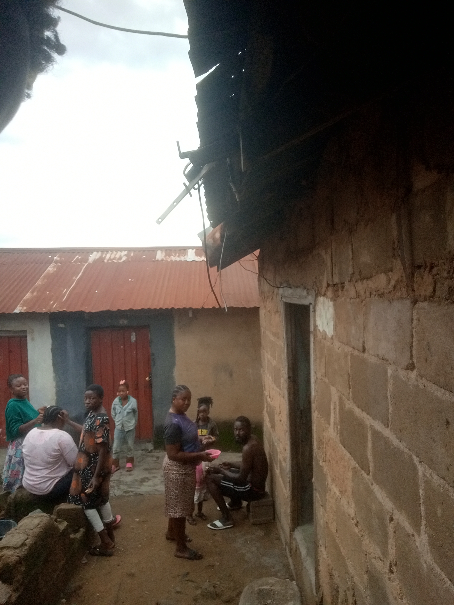 An outdoor scene in Jos, Nigeria, depicts a group of people gathered near a building with a corrugated metal roof. In the foreground, several children and adults are present. A woman in a green top and patterned skirt is attending to the hair of a woman in a pink top and patterned skirt. A girl in a black and orange patterned dress stands nearby. Further back, a person in a light blue shirt and grey pants stands in front of a red door. To the right, a woman in a purple top and patterned skirt holds a pink bowl. Another child is partially visible behind her. A man with a beard, wearing black and white striped shorts, is seated on a small stool against a cinder block wall on the right side of the image. The ground is dirt and appears damp. The building to the right is constructed of cinder blocks with rough mortar. The scene appears to be a residential area.