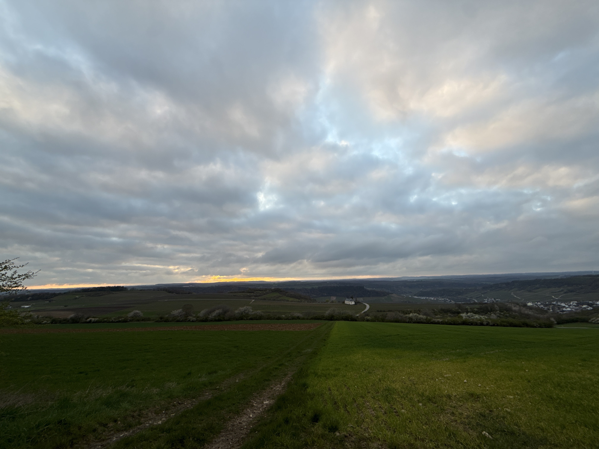 The image depicts a landscape with a large, cloudy sky dominating the upper portion. The sky consists of gray and white clouds, with a hint of light suggesting either sunrise or sunset on the horizon. Below the sky is a panorama of rolling hills and fields, including a green field in the foreground with a dirt path running through it. Further back, there is a line of trees, followed by a mix of cultivated land and natural vegetation. A small building with a light-colored roof is visible in the middle distance. A town or settlement can be discerned in the far distance. The location is unknown.