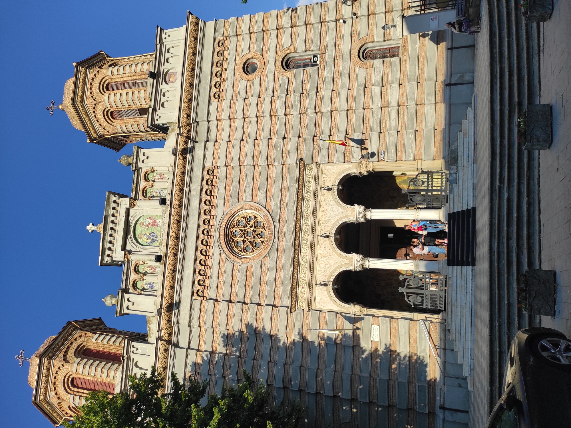The image shows the exterior of a large, ornate church, likely an Orthodox cathedral given the architectural style and the presence of mosaics depicting religious figures. The building features two prominent bell towers with golden domes, brickwork, and arched windows. The facade is made of stone and brick, with decorative elements and a large circular window above the entrance. Several people are visible on the steps leading up to the main entrance, some of whom appear to be entering or exiting the building. A Romanian flag is displayed on the side of the building. In the foreground, a portion of a dark-colored car is visible. The scene is set against a clear blue sky, suggesting it is daytime. The style of the church is characteristic of Romanian Orthodox cathedrals, and specifically, the Metropolitan Cathedral of Saints Peter and Paul in Constanța, Romania, matches the architectural details seen in the image, including the twin towers, domes, and facade design.