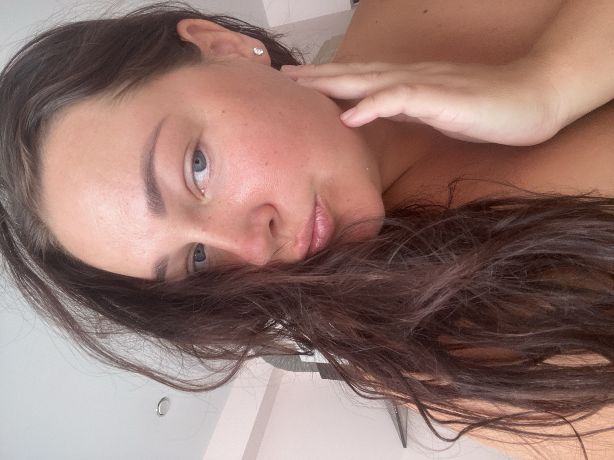 A close-up, eye-level selfie of a woman with long, dark brown hair pulled to the left side of her face. Her skin appears flushed with some redness on her cheeks. She is wearing a small, clear stud earring in her right ear. Her lips are pursed, and her right hand is gently touching her jawline. Her eyes are open and looking towards the camera. The background is indistinct, appearing to be an indoor setting with a plain, light-colored wall and a ceiling light visible in the upper left quadrant. There is no indication of the city of Sochi, Russia, in the image.