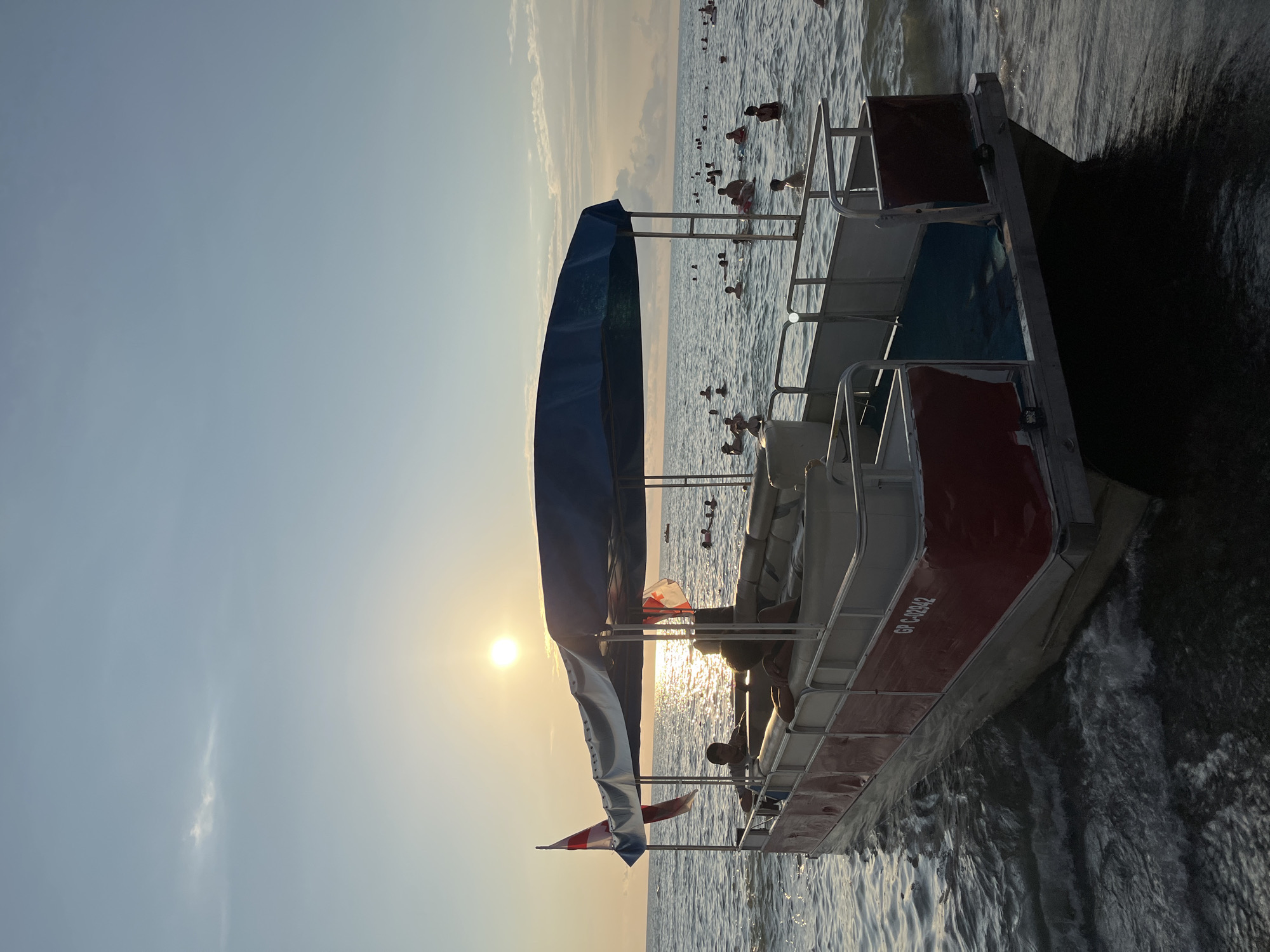 In the city of Ureki, Georgia, at sunset, a pontoon boat is positioned in shallow water near the shore. The boat has a blue canopy and is red and white in color. Two individuals are seated on the boat, with two flags on poles at the front. The sun is setting over the sea, casting a golden glow on the water. Numerous people are swimming and wading in the sea in the background. The boat has the registration number "GP C-02942" visible on its side. The sea is calm with small waves.