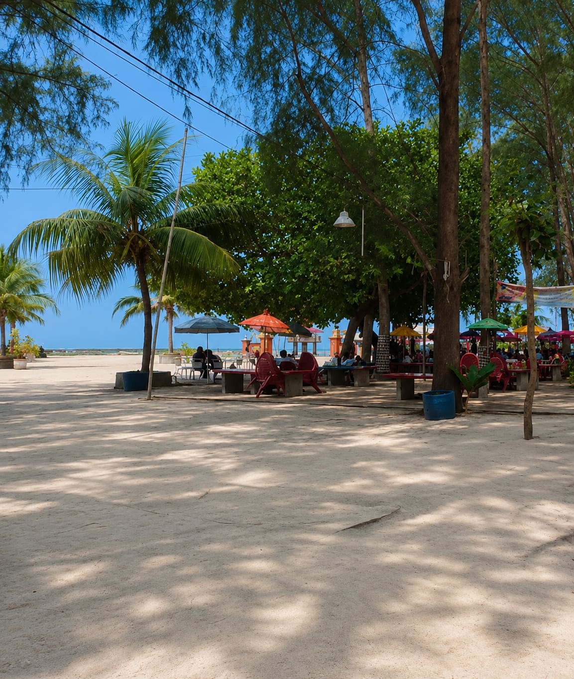 This image depicts a beachside seating area with several people. The scene is set in Kota Pari, Indonesia, characterized by its sandy terrain and tropical vegetation. In the foreground, a wide expanse of light brown sand is dappled with shadows from overhead trees, suggesting a sunny day. Several tables and chairs, mostly red, are arranged in small clusters, some with umbrellas providing shade.
Several individuals are seated at these tables, engaged in conversation or relaxation. They appear to be patrons of a beachside establishment. Behind the seating area, lush green trees form a dense canopy, with palm trees also visible, contributing to the tropical atmosphere. The background reveals a clear blue sky and the distant ocean. Several colorful umbrellas are spread out, indicating a functional outdoor dining or lounge space. A blue trash bin is visible near one of the tables. Power lines stretch across the upper portion of the image, indicating the presence of infrastructure. The overall impression is one of a casual, relaxed beachside environment.