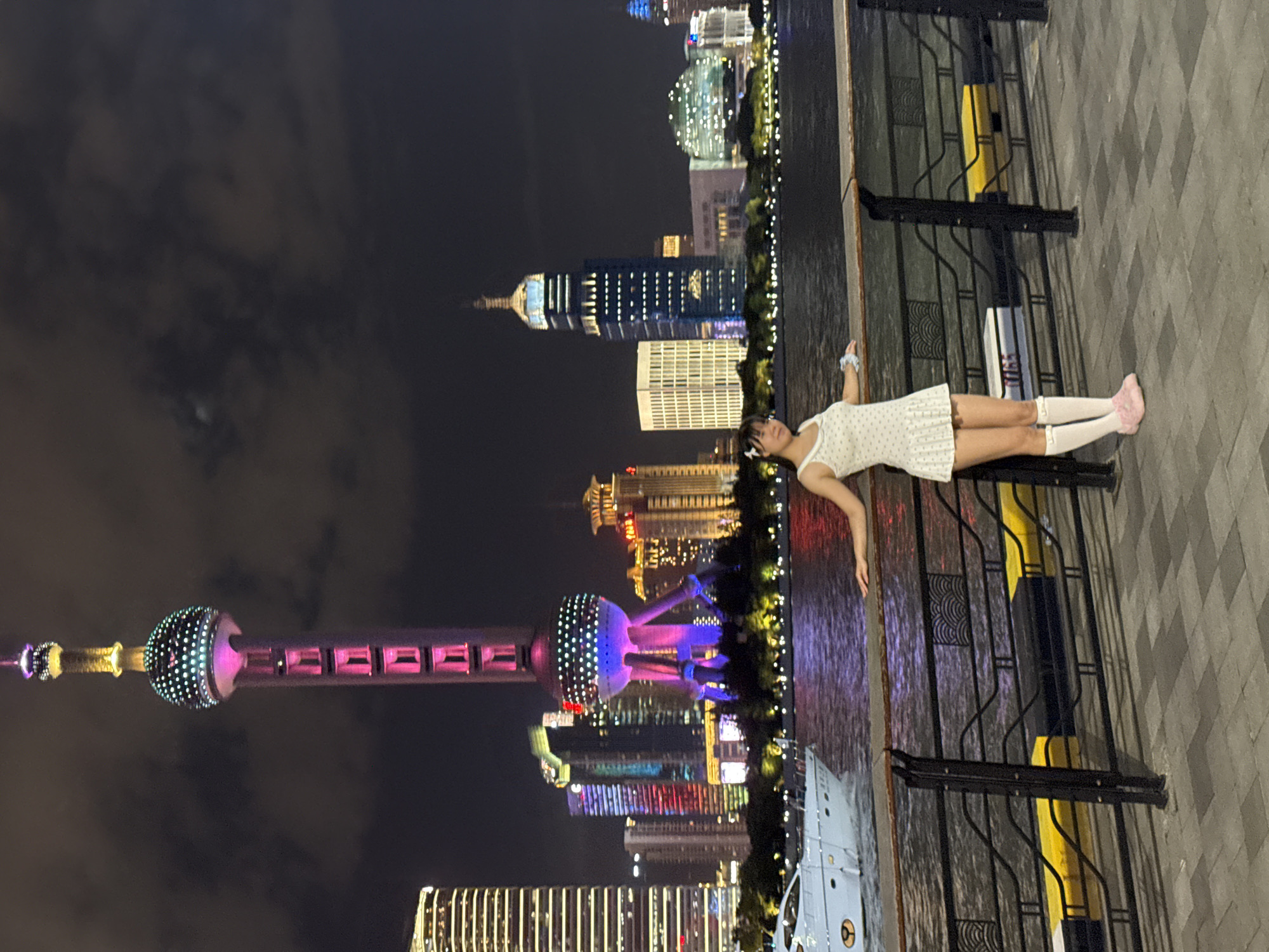In the Hongkou District of China, a young woman stands by a railing at night. She is wearing a white polka-dot dress, white knee-high socks, and pink slip-on shoes. Her hair is adorned with small white flowers. Behind her, the Oriental Pearl Tower, illuminated with pink and purple lights, dominates the skyline. The surrounding cityscape is a vibrant display of illuminated skyscrapers. A white yacht is visible on the water in the foreground. The scene captures a night view of the Bund waterfront.
