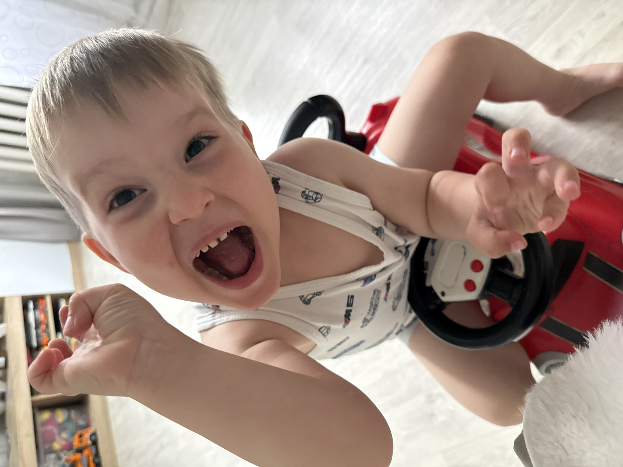 A young Caucasian boy with blonde hair and blue eyes is sitting on a red toy car, which has a black steering wheel with red buttons. He is wearing a white tank top with a pattern of vehicles. His mouth is wide open in a joyful expression, and his left arm is raised with his hand in a fist. The boy's right hand is extended, palm up. The scene appears to be indoors, with a light-colored floor and a blurry background showing shelves with toys and possibly a radiator. The lighting is bright, suggesting daylight.