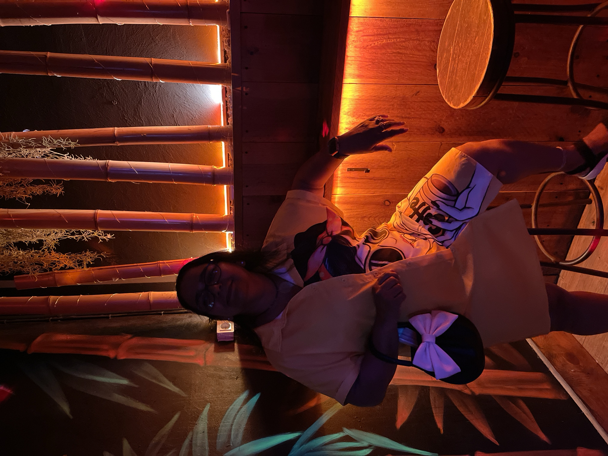A person, wearing glasses and a yellow short-sleeved collared shirt over patterned shorts, sits with their legs extended. They are holding a black purse with a large white bow on it. The person has dark hair and appears to be smiling faintly. The background is dimly lit with warm, orange-toned lighting. Visible elements of the setting include bamboo stalks arranged vertically, some with lights behind them, and wooden paneling. A circular wooden table top is visible above and to the right of the person. The overall scene evokes a tropical or tiki-themed bar or restaurant. The wall also features painted green bamboo leaves. The image is oriented sideways.