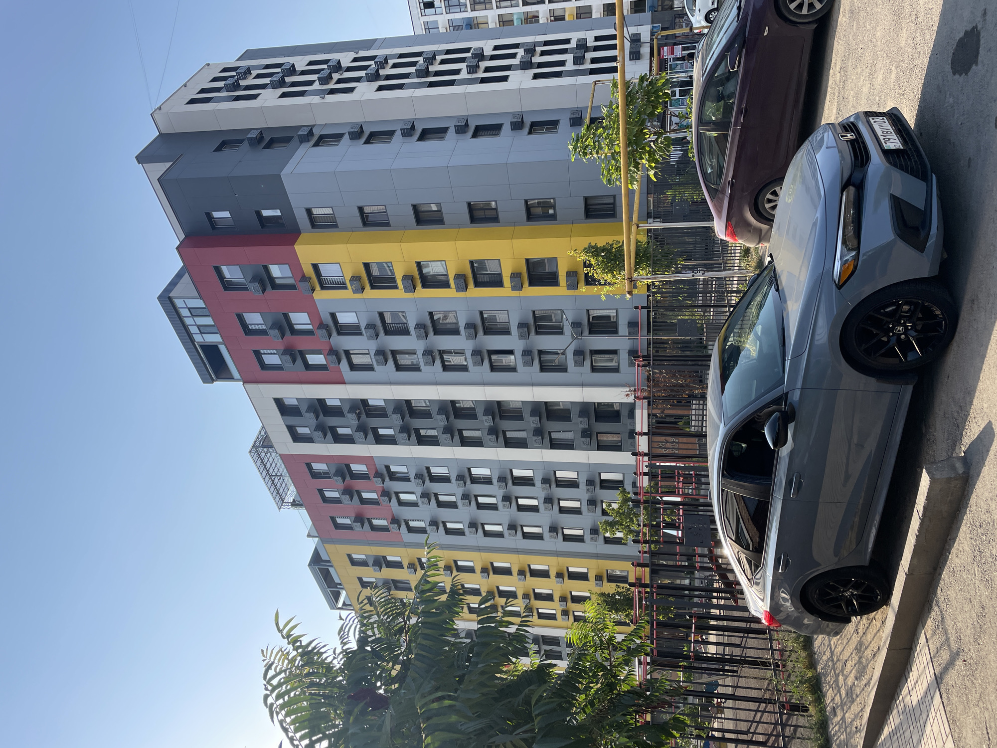 In Almaty, Kazakhstan, on a clear day, a modern, multi-colored apartment building stands tall. The building's facade is segmented into vertical bands of red, yellow, and varying shades of gray and white, with numerous windows and external air conditioning units visible. In the foreground, two cars are parked on a paved area bordered by a sidewalk and a concrete curb. The car closest to the viewer is a gray sedan with black rims. Behind it, a dark purple sedan is also parked. A black metal fence runs along the property line, separating it from a strip of greenery and a walkway or play area. Trees and foliage are present on the left side of the image, partially obscuring the view of the building. The sky is a clear, pale blue.