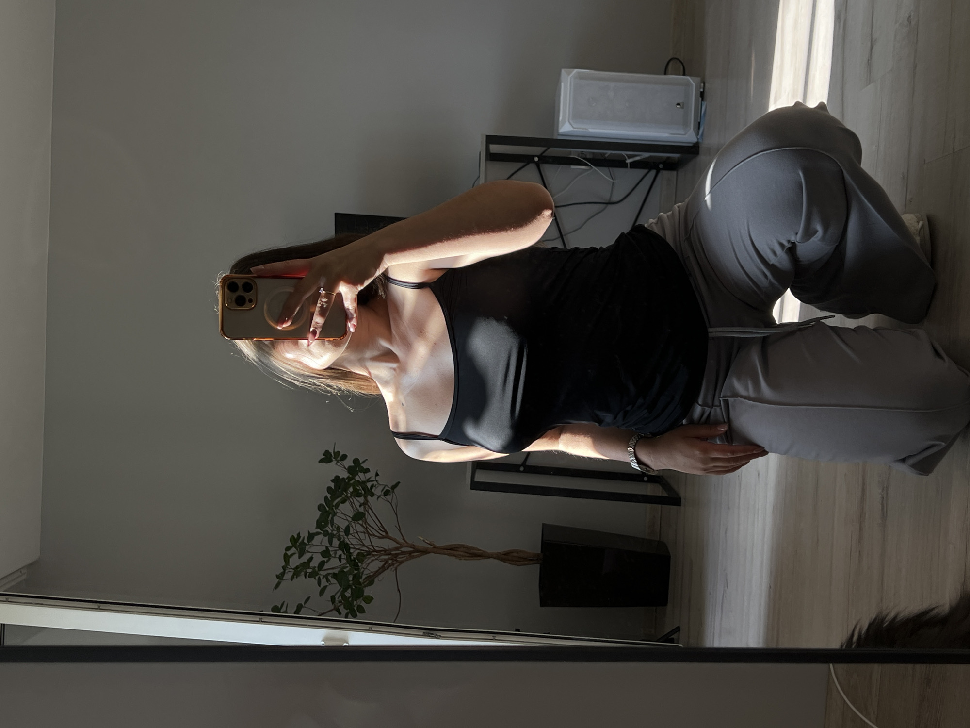 A young woman is kneeling on a wooden floor, taking a selfie in a full-length mirror. She is wearing a black tank top and grey sweatpants. The room is lit by natural sunlight streaming in from the right, creating dramatic shadows. In the background, a tall potted plant sits in a black planter, and a metal shelf unit holds a white computer tower and some cables. The overall environment suggests a domestic setting, possibly in an apartment in Ternopil, Ukraine.