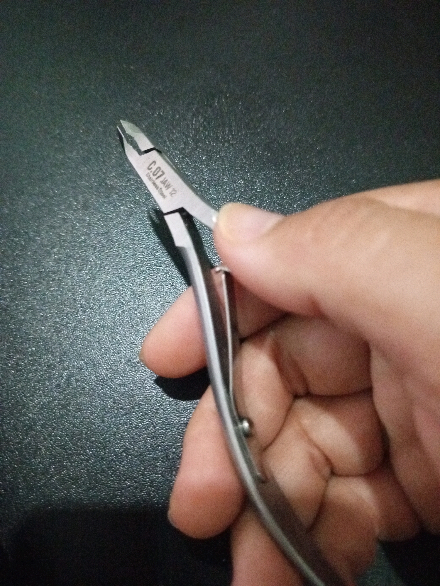 A close-up shot shows a person's hand holding a pair of cuticle nippers. The nippers are made of stainless steel and have a model number "C.07 JAW 12" engraved on them. The background is a dark, textured surface. The image was taken in Denpasar, Indonesia.