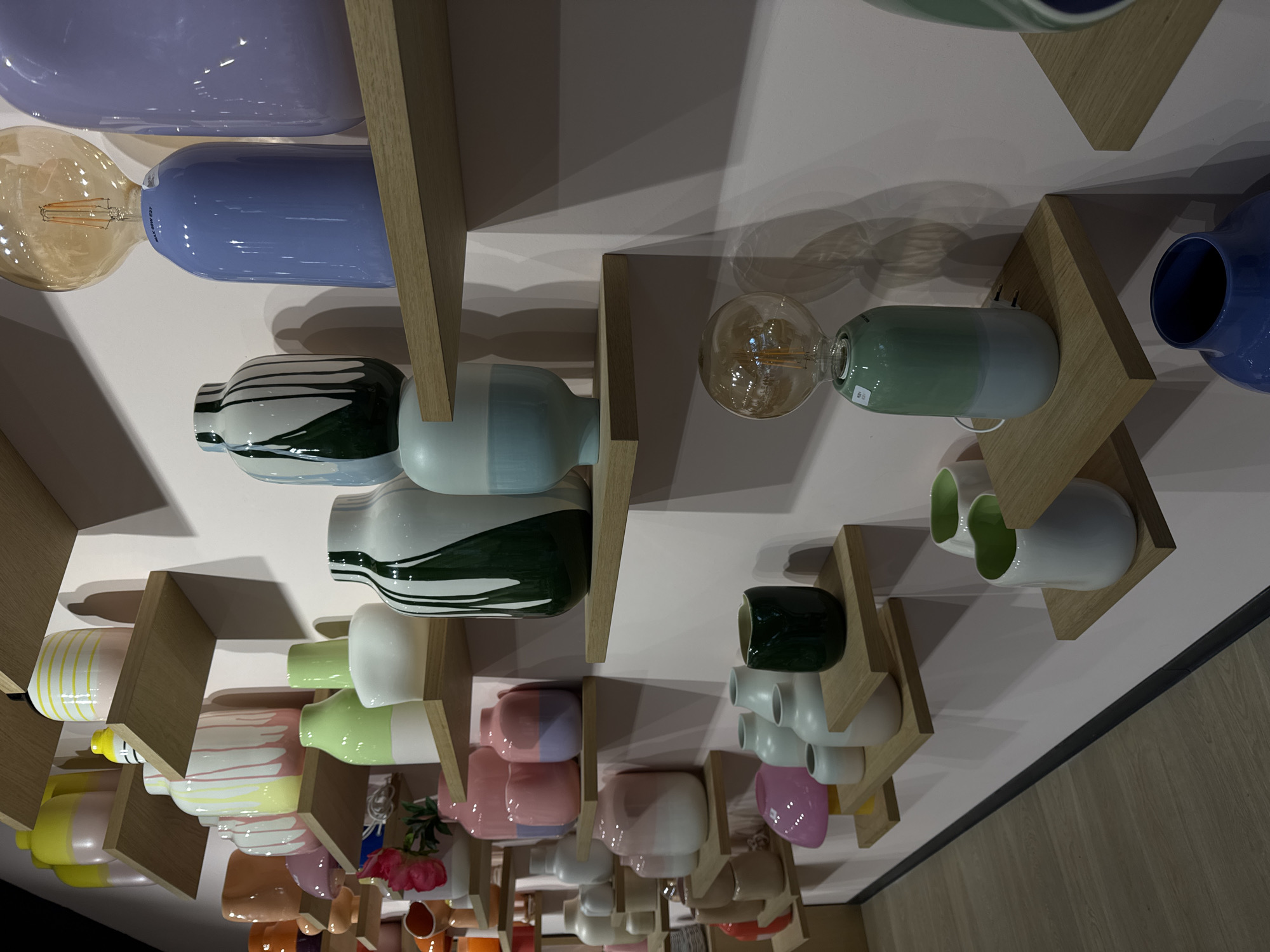 The image shows a display of colorful vases and light bulbs on wooden shelves, presumably in a retail setting. The shelves are mounted on a light-colored wall. The vases are various shapes and colors, including white, green, blue, pink, yellow, and orange, with some featuring patterns. Some light bulbs are attached to some of the vases.
Although the request mentions Roermond, Netherlands, there is no explicit evidence within the image itself to confirm the location. Therefore, I cannot definitively determine if the image was taken in Roermond.