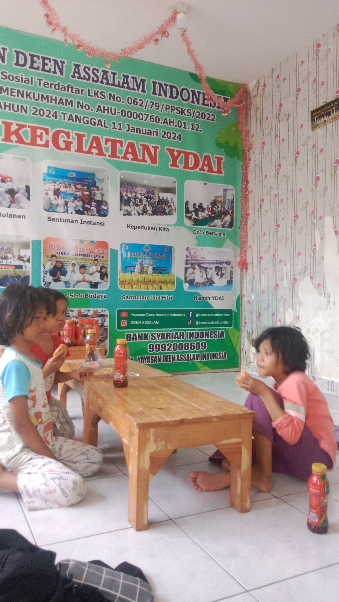 In a room with tiled flooring and patterned wallpaper, a wooden table sits in the foreground. Three young children are gathered around the table, which holds several bottles of drinks. The child on the left, a girl wearing a blue and white pajama set, is looking down at the table. Next to her, another girl in a red shirt is holding a piece of food in her hand. The child on the right, a girl in a pink shirt and purple pants, is looking to her right and eating. Behind the children, a large banner is prominently displayed, featuring text in Indonesian and several smaller images depicting various activities. The banner text indicates that this is an event for "Yayasan Deen Assalam Indonesia" and mentions "KEGIATAN YDAI" and "BANK SYARIAH INDONESIA". A detail on the banner reads "DESA SUMBER JAYA", indicating the location as Sumber Jaya Village, Indonesia. The overall scene appears to be an indoor gathering or event.