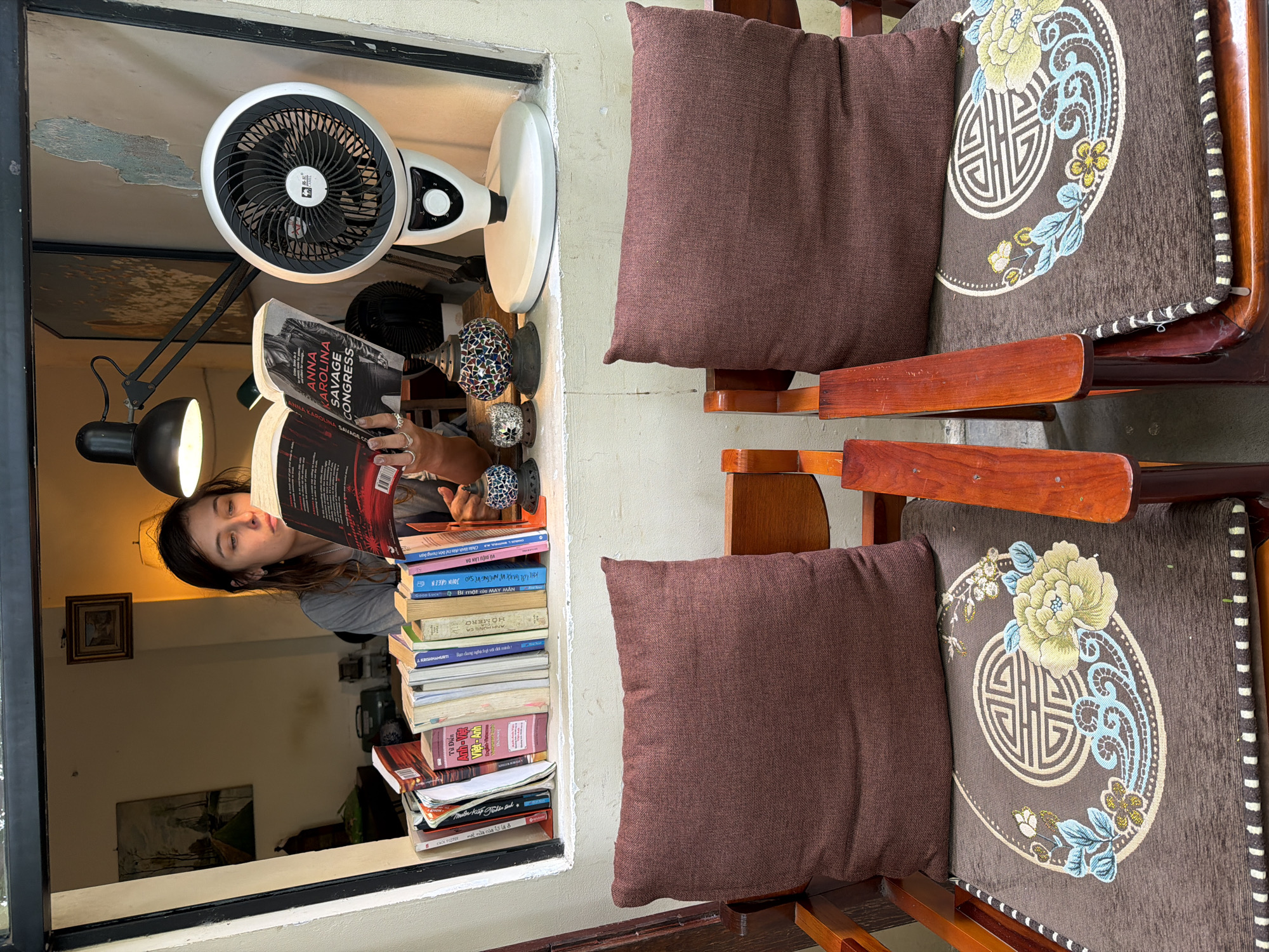 The image depicts an indoor scene, likely in Đà Nẵng, Vietnam. A young woman with brown hair is seated at a window, engrossed in reading a book. She is wearing a grey t-shirt and has several rings on her fingers. The book she is reading has a red and black cover with text that is partially visible.
In front of the woman, on a shelf or windowsill, is a collection of books of various sizes and colors. Behind the books, a desk lamp with a black metal arm is positioned, casting light on the reading area. To the right of the lamp, a white electric fan with a black grill is visible. The window area itself has a slightly distressed, textured wall.
In the foreground, two wooden chairs are positioned. Each chair has a dark brown cushion on the seat and a slightly darker brown pillow at the back. The seat cushions are adorned with a circular design featuring a floral motif and a central geometric pattern. The chairs appear to be made of polished wood.
The overall impression is a cozy and studious atmosphere, possibly within a home or a small shop.