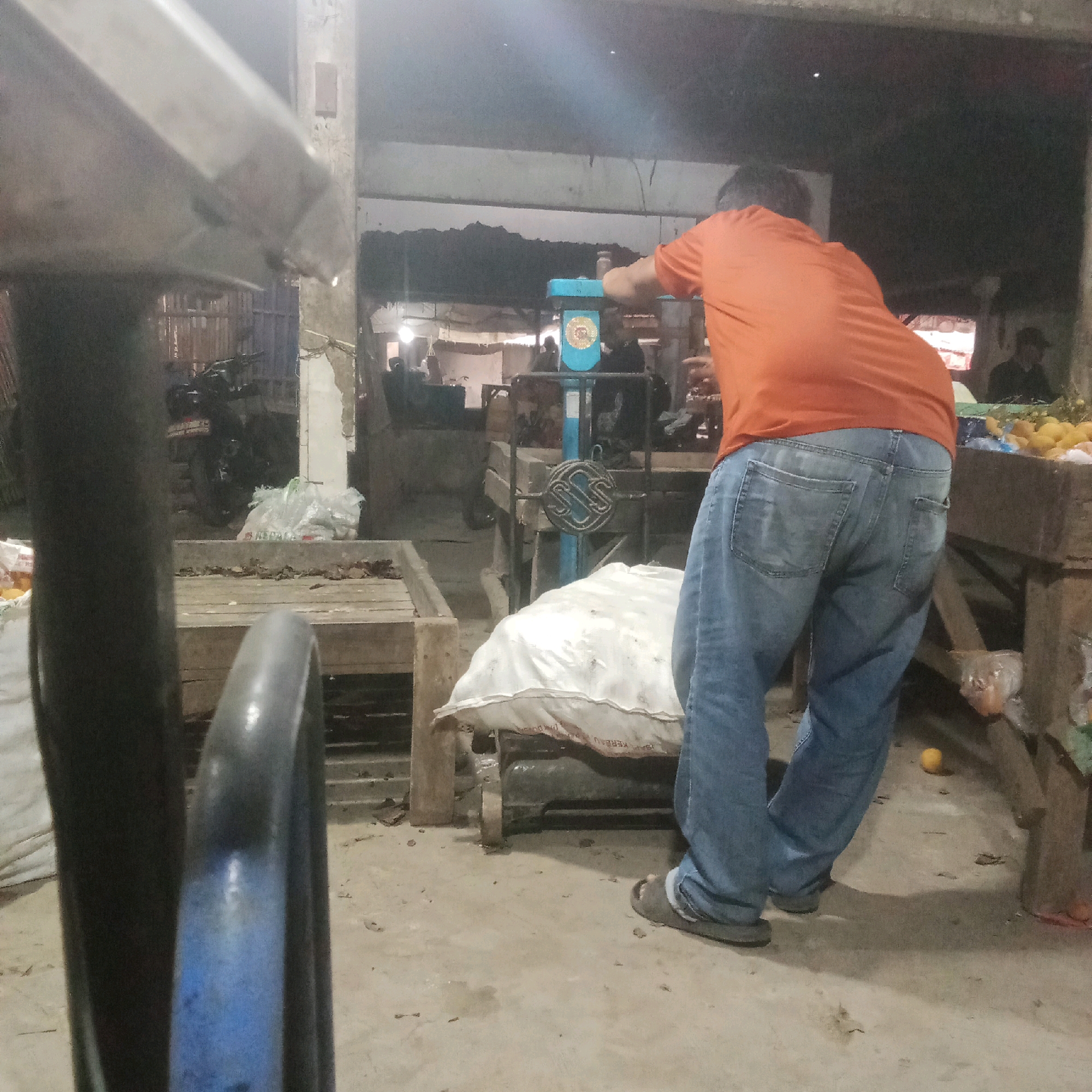 In Karawang, Indonesia, a person wearing an orange t-shirt and blue jeans is standing with their back to the camera, leaning on a blue and white weighing scale. The scale has a circular, decorative element on its front. To the left of the scale is a large, white sack. In the background, there are wooden structures, various items on tables, and people blurred in the distance. A motorcycle is visible to the left of the frame, and several plastic bags are scattered on the ground, some containing produce like oranges. The ground is concrete and appears somewhat dirty, with small debris present. The lighting suggests an indoor or covered market environment.