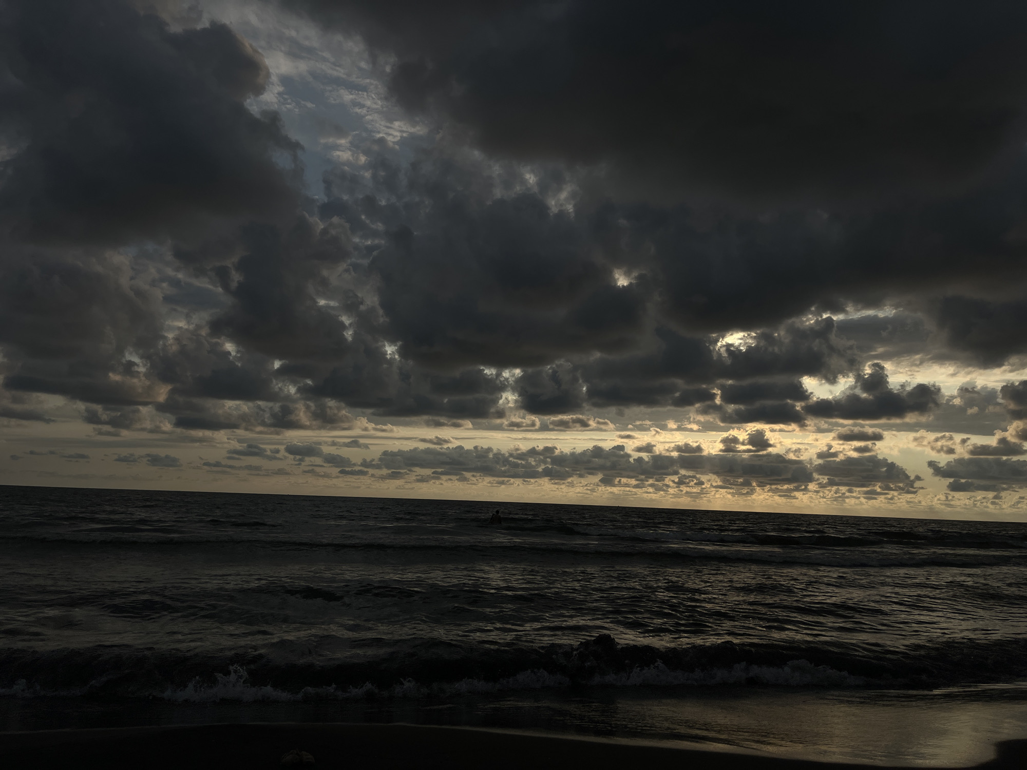 This media file captures a dramatic seascape under a stormy sky. The scene is set at a beach, likely by the Black Sea given the location context of the Supsa community in Georgia. The time of day appears to be late afternoon or early evening, as evidenced by the low angle of the sun peeking through the dense, dark clouds, creating a moody and atmospheric light. The weather is clearly turbulent, with heavy, overcast clouds dominating the upper portion of the frame.
The ocean is a dark, choppy expanse, with waves gently breaking on the shore, creating white foam. There are faint ripples on the water's surface, suggesting a moderate breeze. In the distance, a solitary figure is visible in the water, possibly swimming or wading, adding a human element to the vastness of the sea and sky. The beach itself is dark and indistinct in the foreground, with a hint of sand and perhaps some debris.
The overall mood of the image is one of powerful natural forces at play. The dark, brooding clouds and the turbulent sea evoke a sense of awe and perhaps a touch of melancholy. The contrast between the dark clouds and the occasional glimpses of golden light breaking through suggests a dramatic shift in the weather, perhaps the end of a storm or the prelude to one. There is no visible text in the media file.
