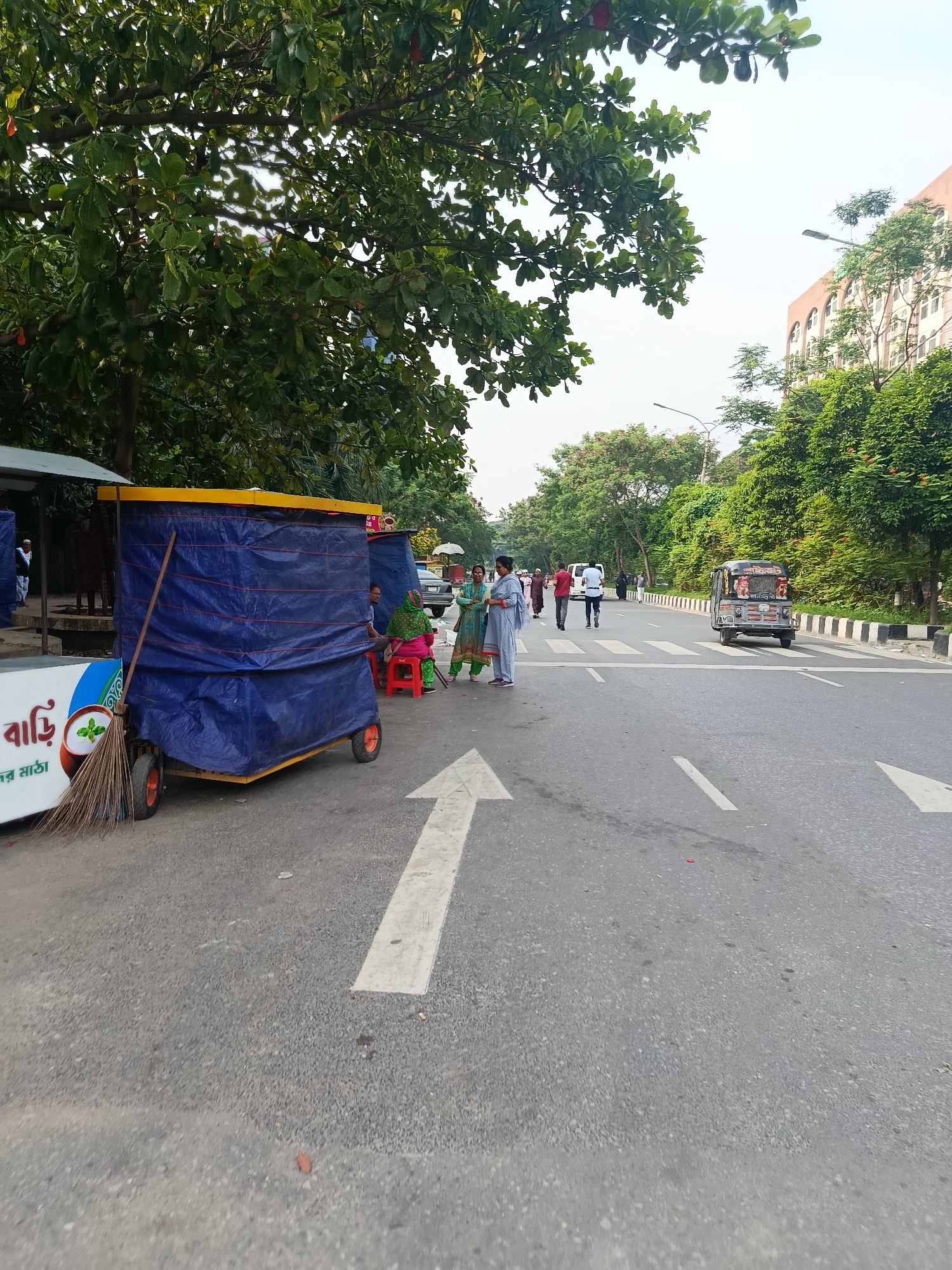 The image depicts a street scene in Dhaka, Bangladesh. A mobile food stall, covered in a blue tarp and advertising a yogurt drink, sits on the side of a road with a large arrow painted on its surface. A woman in a green and pink sari sits on a red stool near the stall. Several people are walking across a pedestrian crosswalk. A small motorized three-wheeled vehicle is driving on the road. The scene is bordered by lush trees and foliage. A multi-story building is visible in the background on the right.