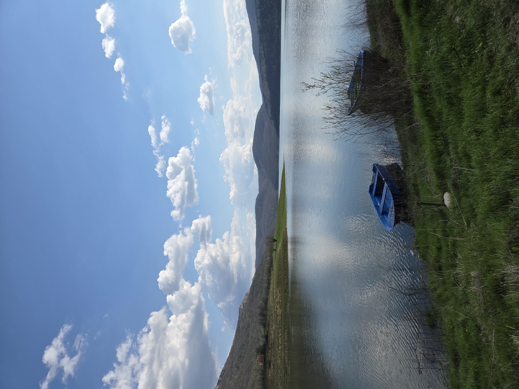 The image captures a serene landscape likely near the city of Debelets, Bulgaria. A body of water, possibly a lake or a wide river, dominates the right half of the frame, reflecting the sky and the mountain ridge present in the middle. Two small blue boats are visible along the bank, nestled amongst the green grass and vegetation. The left half of the image showcases a bright blue sky filled with fluffy white cumulus clouds. A mountain range forms the backdrop, with the landscape transitioning from green at the base to rocky terrain further up. The image suggests a tranquil environment.
