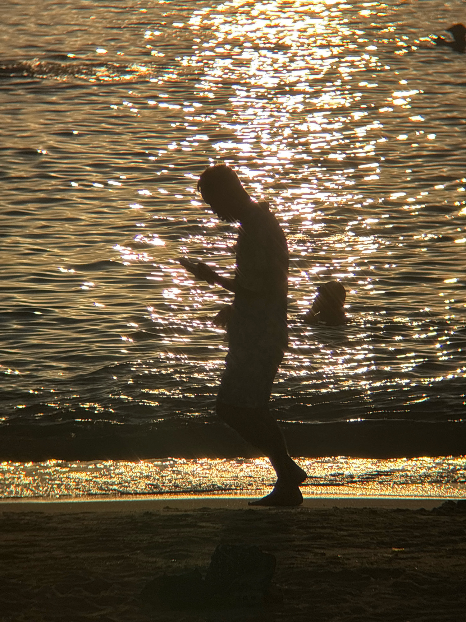 The image is a silhouette of a person standing in shallow water at a beach, likely in Nha Trang, Vietnam, during sunset. The sun reflects brightly off the water's surface, creating a shimmering effect. The person is facing away from the camera, looking down at a mobile device held in their hands. Their body is in profile, with their legs slightly bent as if they are walking or standing still. The water laps at their ankles and calves. Another person, with only their head visible, is in the background, further out in the water. The foreground shows a sandy beach, also in silhouette, with a few dark, indistinct objects that could be rocks or driftwood. The lighting and the silhouettes suggest a warm, evening atmosphere.