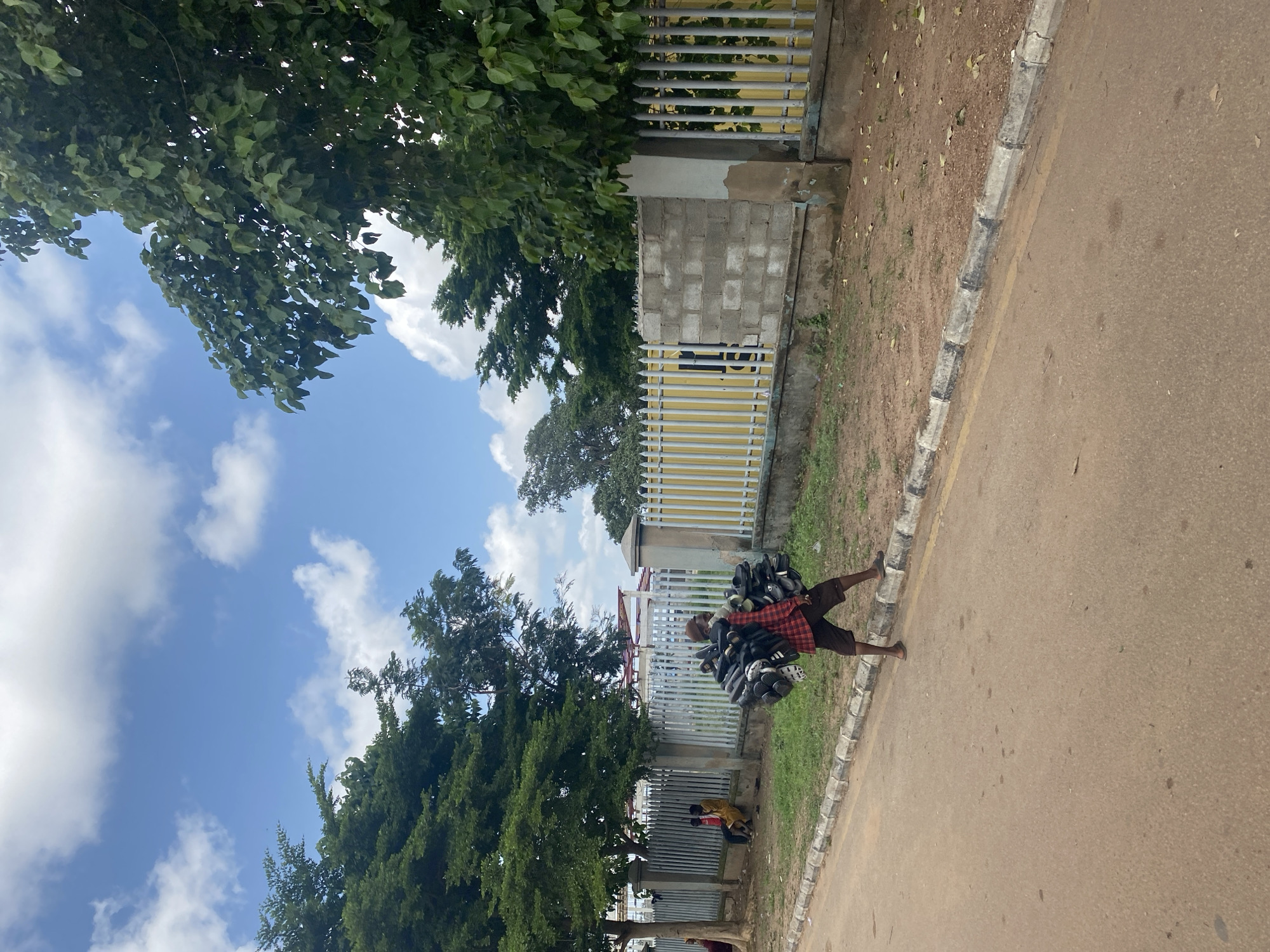 In Ado, Nigeria, a man walks along a dusty road carrying a large bundle of black shoes strapped together. He is wearing a red and black checkered shirt, brown shorts, and sandals. The man is walking on the right side of the road, and to his left is a grey concrete and metal fence with a yellow structure behind it. Trees line the other side of the road and the area behind the fence. In the background, two young boys are sitting near a tree next to the fence. The sky is blue with scattered white clouds.