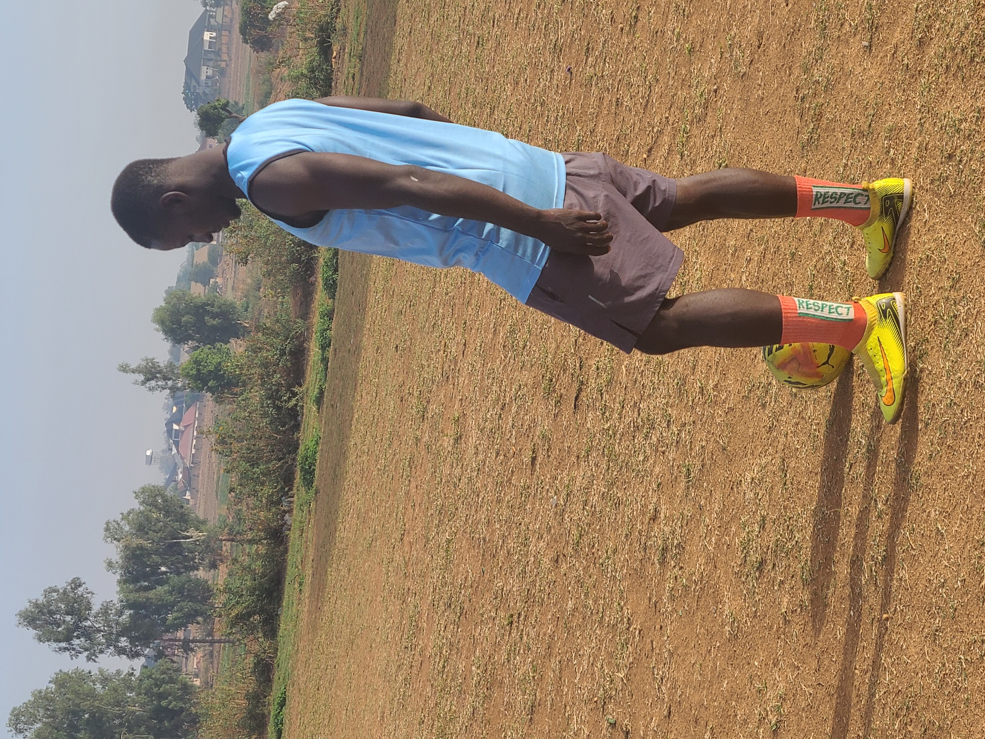 The image captures a dark-skinned man standing on a dry, earthy field in Barikin Jirga Summa, Nigeria, under a bright, clear sky. He is dressed in a light blue sleeveless athletic top and dark shorts. His lower legs are covered by vibrant orange socks, each boldly displaying the word "RESPECT" in green letters. He wears bright neon yellow soccer cleats. One foot rests atop a soccer ball, suggesting he is preparing to play or is posing with it.
The man is seen in profile, looking downwards towards the ball, with his body slightly bent forward in an athletic stance. His arms are relaxed, with the left arm slightly bent at the elbow. Strong, elongated shadows of the man are cast on the ground, indicating bright daylight, likely midday or early afternoon. The field itself is parched, with sparse, dry grass and visible soil. In the background, scattered trees and several distant buildings under a hazy sky provide context to the rural or semi-urban setting. The overall scene suggests a sunny, dry day at a local playing field.