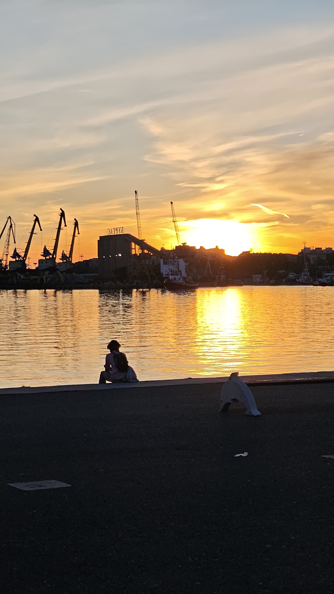 The image shows a sunset scene in the city of Burgas, Bulgaria. The location appears to be a waterfront area or harbor, with cranes and industrial buildings visible in the background. A person is sitting on the edge of the waterfront, facing the water and watching the sunset. The sun is setting over the water, creating a golden reflection on the surface. In the foreground, there is a plastic dolphin-shaped barrier and some litter on the ground.