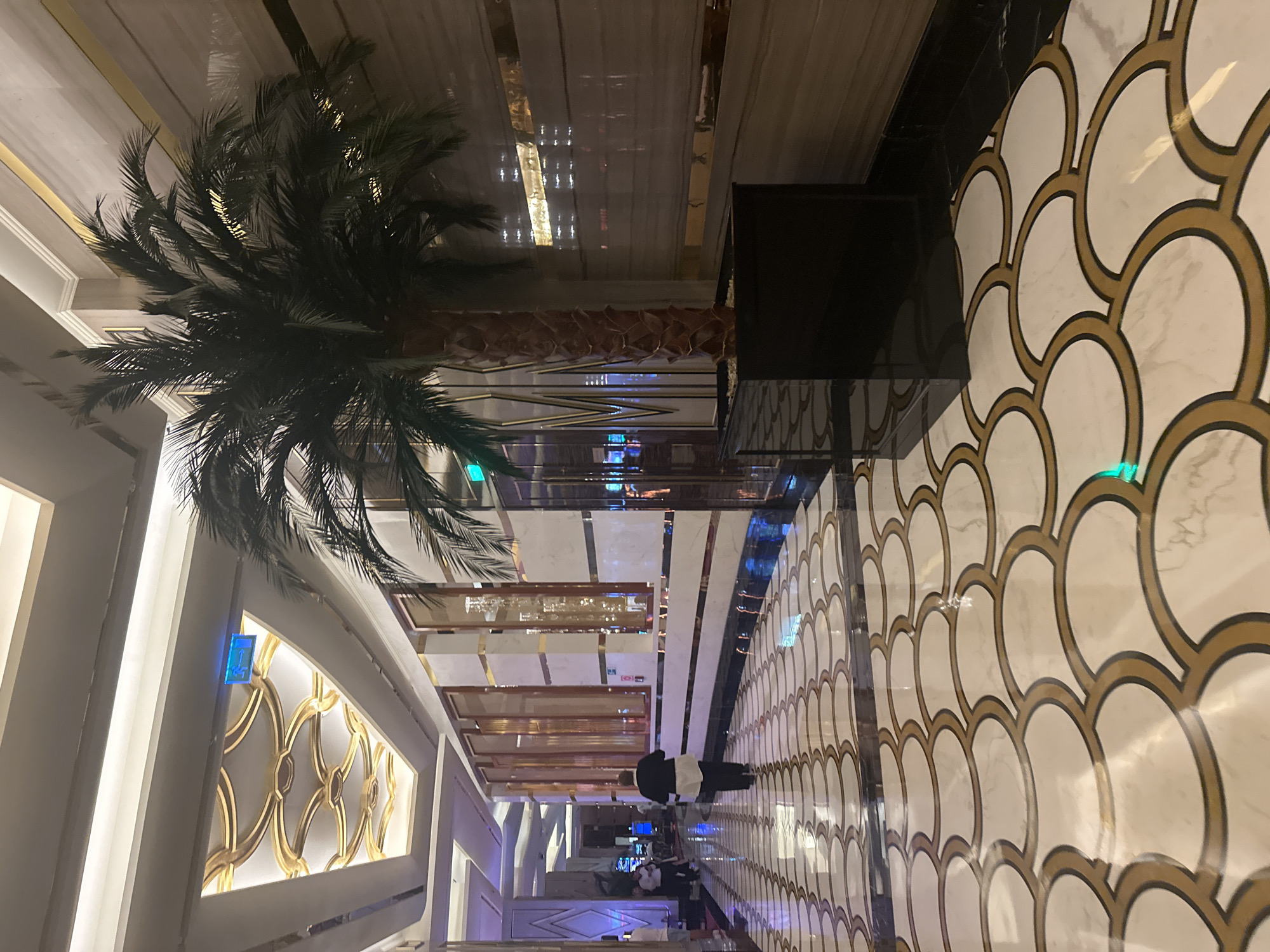 The image shows a wide, ornate hallway with a highly polished marble floor featuring a repeating pattern of white and gold semi-circular shapes. On the right side of the hallway, a tall artificial palm tree in a black planter is visible, with its fronds extending towards the ceiling. The walls are clad in a light-colored, textured material with gold horizontal accents. The ceiling is intricately designed with gold, curved elements and recessed lighting. Further down the hallway, several people are walking. One person, dressed in a black top and dark pants, is walking away from the camera. The hallway appears to be an interior space, likely a hotel or casino lobby, given its luxurious design. The location is identified as Estosadok, Russia.