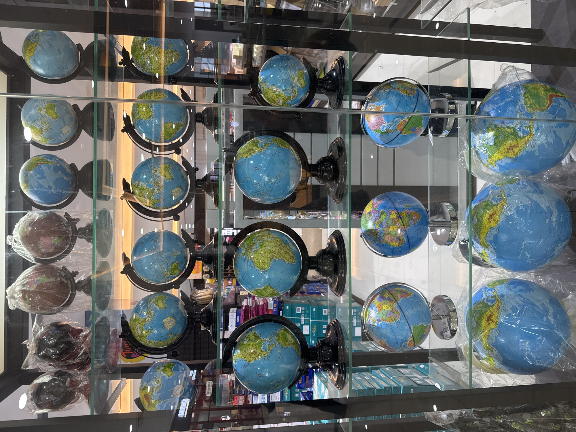 The image displays a retail setting featuring numerous globes showcased on glass shelves within a dark-framed display cabinet. The globes vary in size and style; most have blue oceans with landmasses depicted in shades of green, yellow, and brown, indicating political or physical maps. Many smaller globes are mounted in black, arched stands, while some larger ones are wrapped individually in clear plastic for protection.
The glass shelves produce prominent reflections, mirroring the globes themselves, ceiling lights, and other parts of the store, creating a sense of depth and abundance. Several globes clearly show names of continents and oceans, such as "North America," "South America," "Africa," "Europe," "Asia," "Atlantic Ocean," and "Pacific Ocean."
In the background, blurred by reflections and visible through the glass, are stacked products in boxes and other store items. Faint text such as "CUSTOM," "KLASSIK," "SIDO," and "PARKS" can be seen on labels or packaging. The scene is brightly lit, suggesting an indoor store environment in Kuta, Indonesia, where these globes are available for purchase.