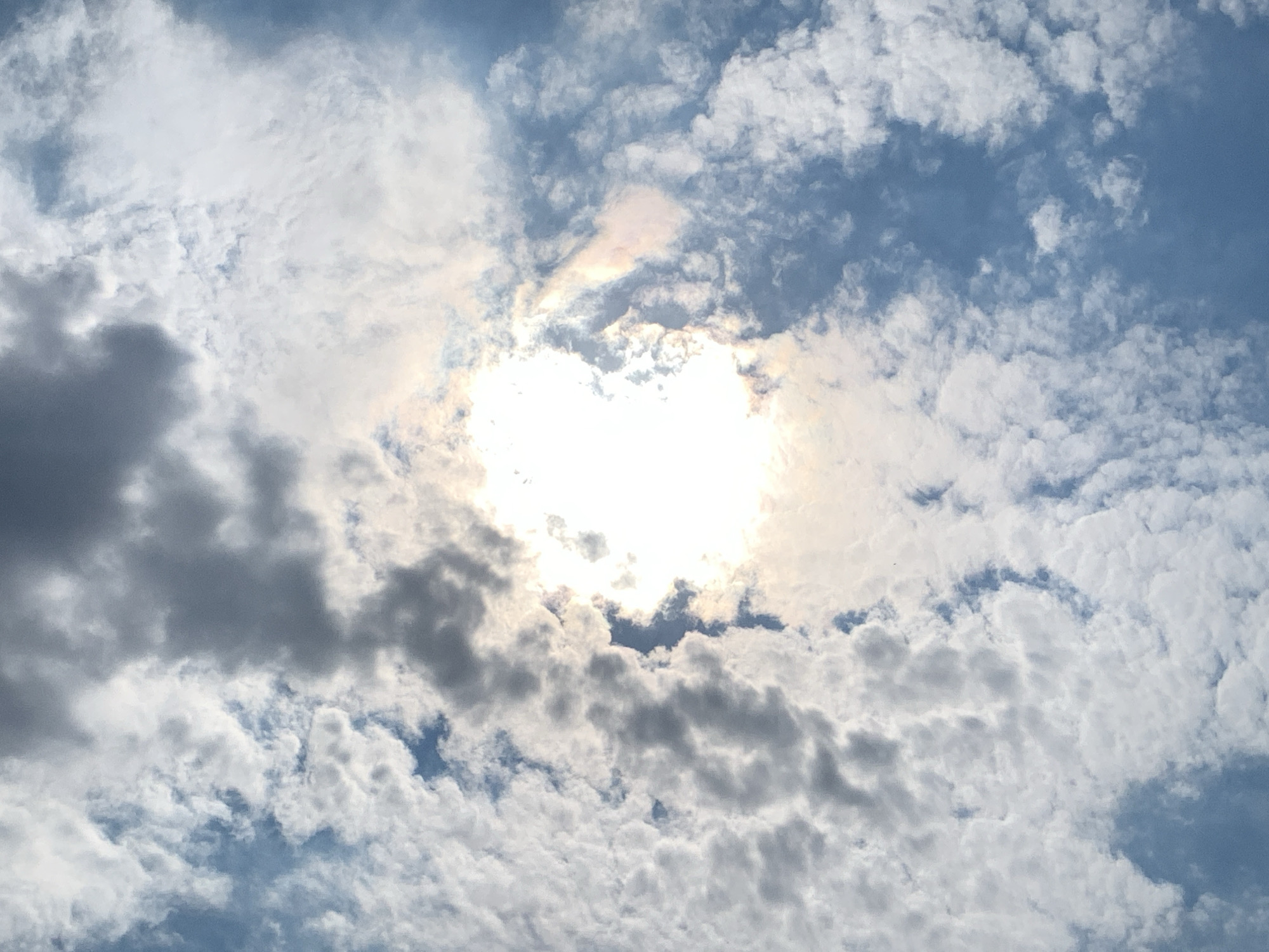 The image shows a sky filled with a mix of white and dark clouds, with the sun shining brightly through a gap in the clouds. The sun's rays create a luminous effect, and there are hints of rainbow colors in the clouds around the sun. The sky is a clear blue in some areas where the clouds are thinner. The scene appears to be an outdoor shot, likely taken from the ground looking up at the sky, with no visible urban elements or people. The weather appears to be partly cloudy with bright sunshine.