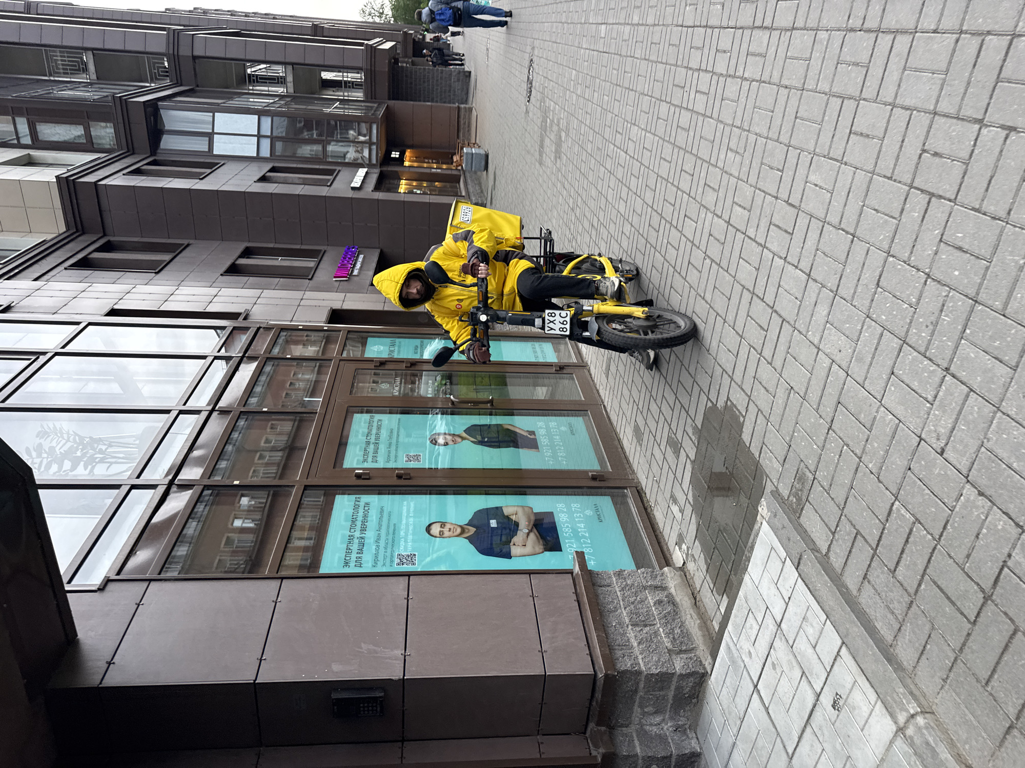 A delivery driver in a bright yellow uniform is seated on a yellow e-bike on a sidewalk in Saint Petersburg, Russia. The bike has a yellow delivery box on the back, and the license plate reads "YX8 86C". The building behind the driver has large glass windows and displays advertisements for a dental clinic called "КРИСТАЛЛ", featuring headshots of doctors and contact information. Pedestrians are visible in the background, walking on the same sidewalk. The street is paved with gray rectangular paving stones.
