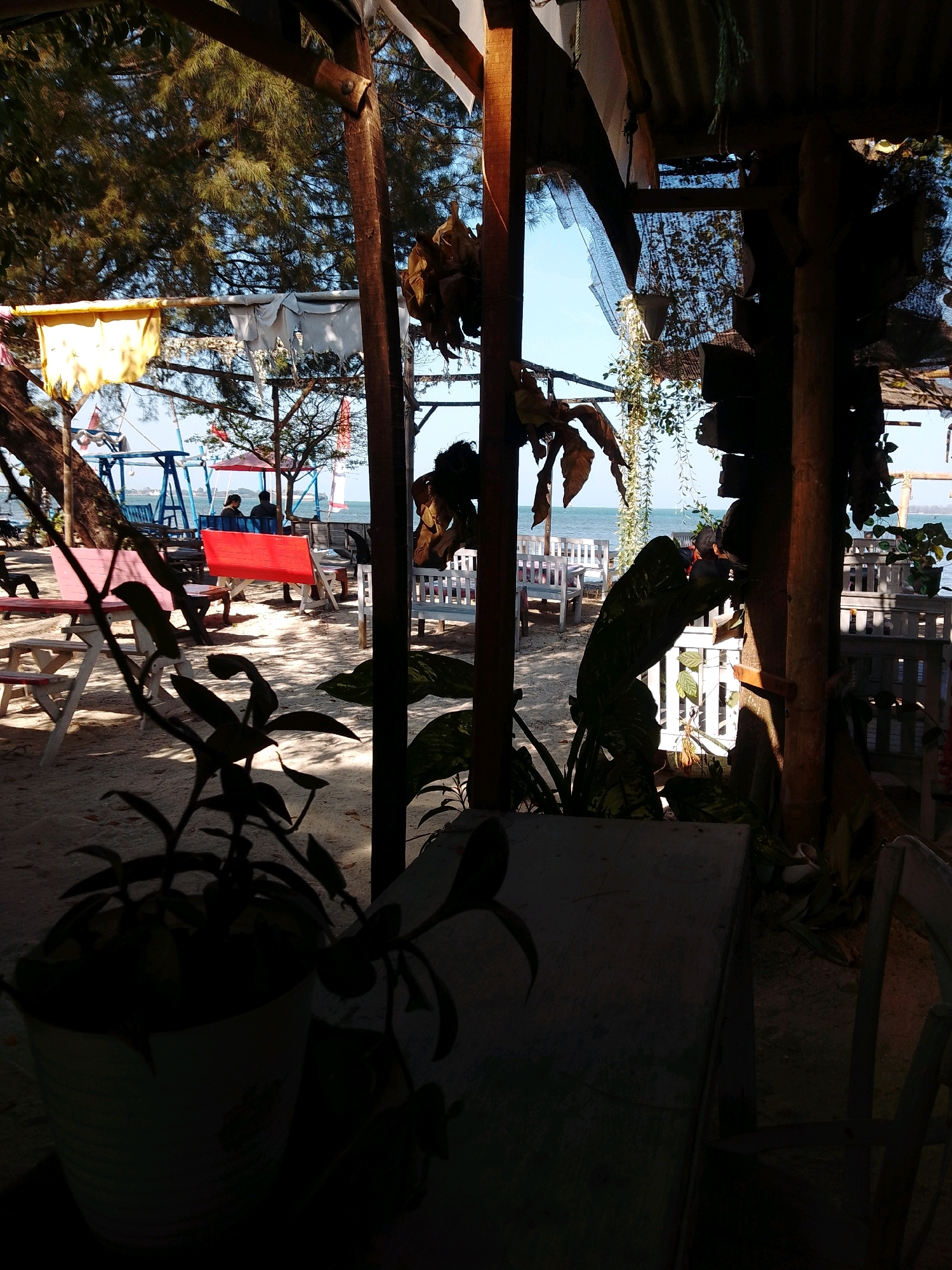 The image depicts an outdoor seating area on a sandy beach, likely at a seaside restaurant or cafe in Cikal, Indonesia. The foreground is dominated by a silhouette of a potted plant and a wooden table, suggesting a viewpoint from within a covered structure. In the mid-ground, several wooden tables and benches are visible, some painted pink and red, arranged on the sand. Two people are seated at a table in the mid-ground, facing away from the camera, with their backs to the sea.
Beyond the seating area, a swing set with blue and red accents is visible. The background features the calm sea under a clear blue sky, with a distant horizon line. The environment is characterized by sandy terrain, sparse vegetation, and overhead structures constructed from wood and corrugated metal, providing shade. The overall impression is a relaxed, beachfront atmosphere.
